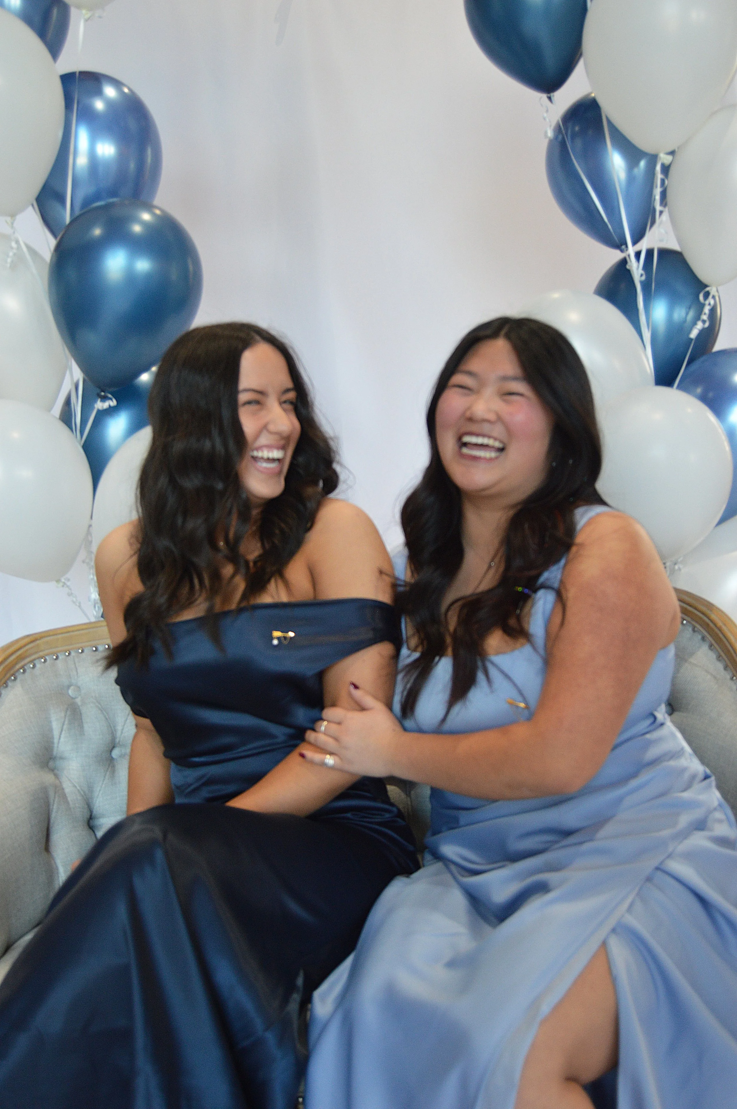Two women sitting on a beige couch, wearing blue dresses, laughing and smiling, surrounded by blue and white balloons.