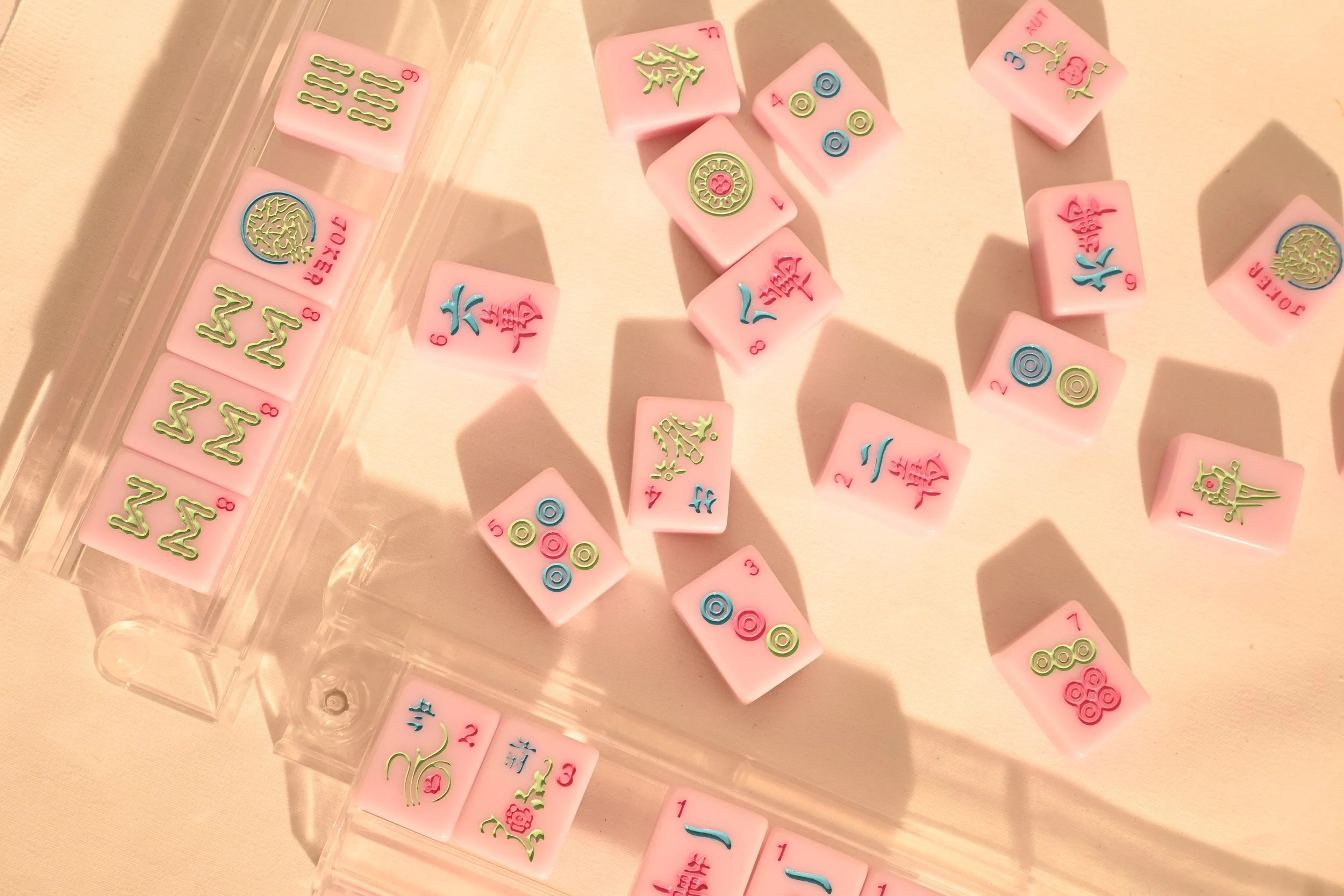 Pink Mahjong tiles with colorful Chinese characters, numbers, and symbols, some inside a clear plastic tray on a beige surface.