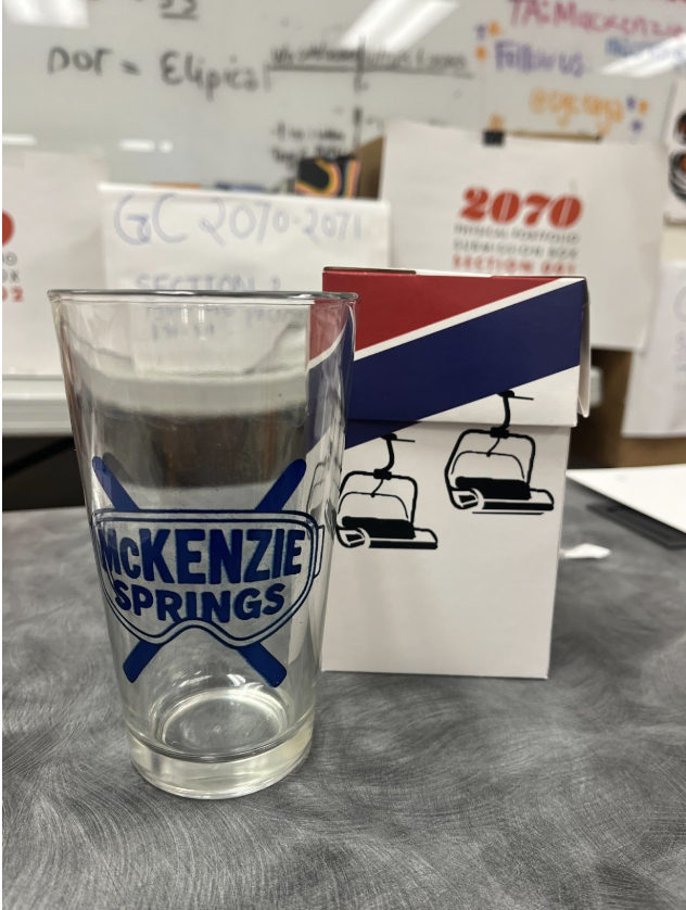 A clear glass with a blue and white 'McKenzie Springs' logo, sitting on a gray table next to a cardboard box with graphics of ski lifts, against a background of whiteboard and boxes.