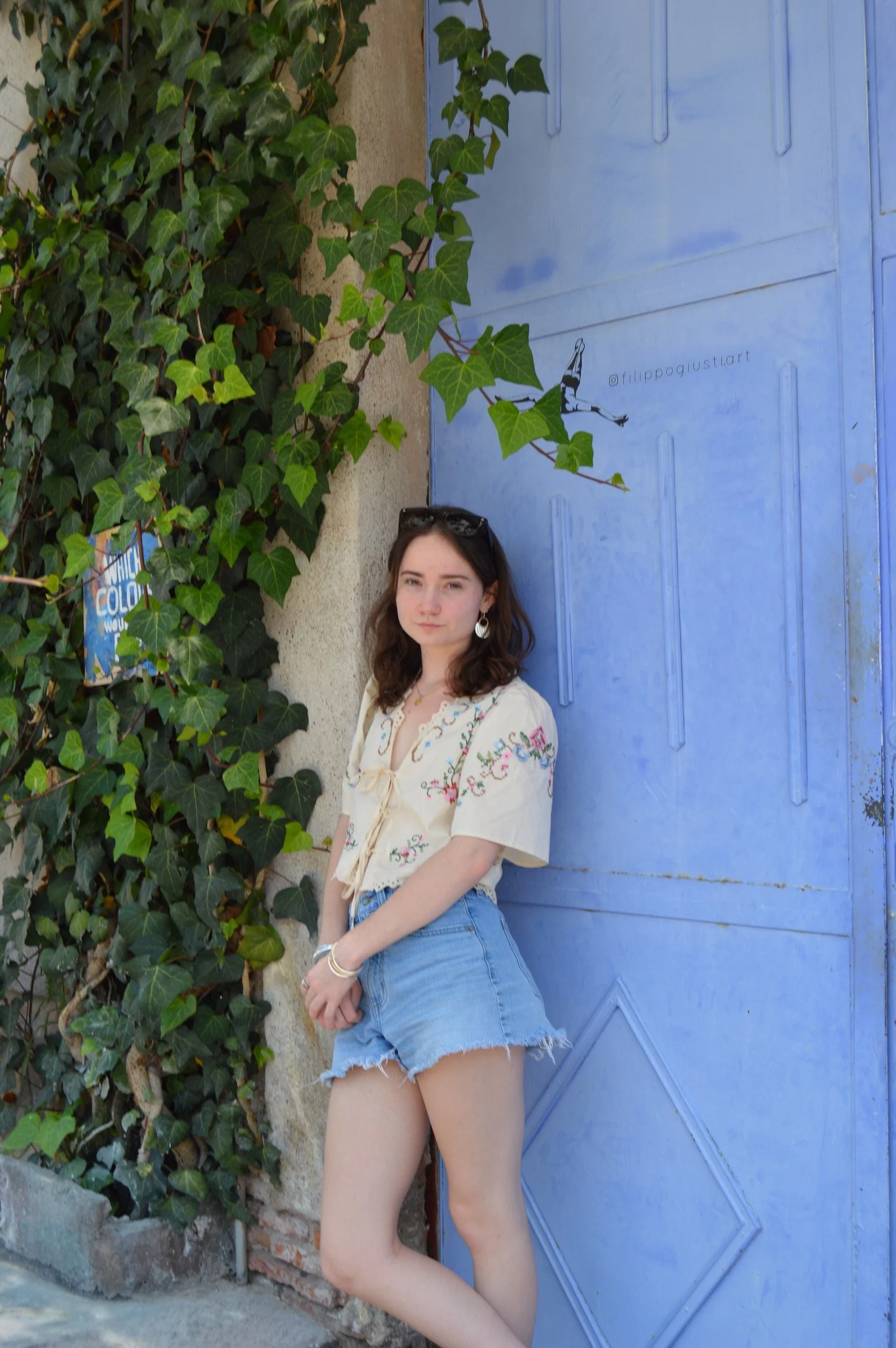 A young woman with brown hair, wearing a cream embroidered blouse and blue denim shorts, stands against a blue door with a vine growing on the wall.