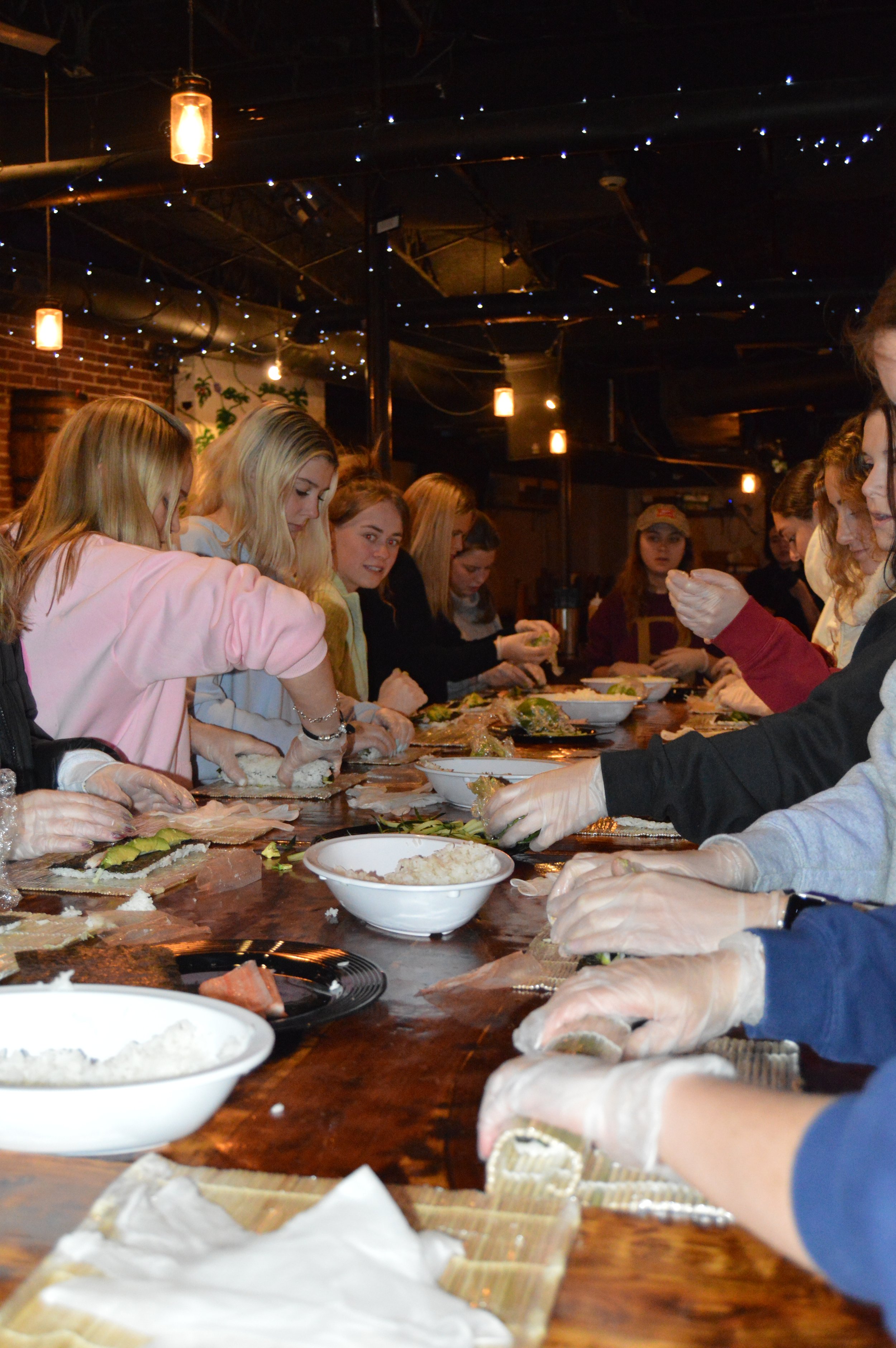 A group of women preparing sushi together at a long table with bowls of rice and other ingredients.