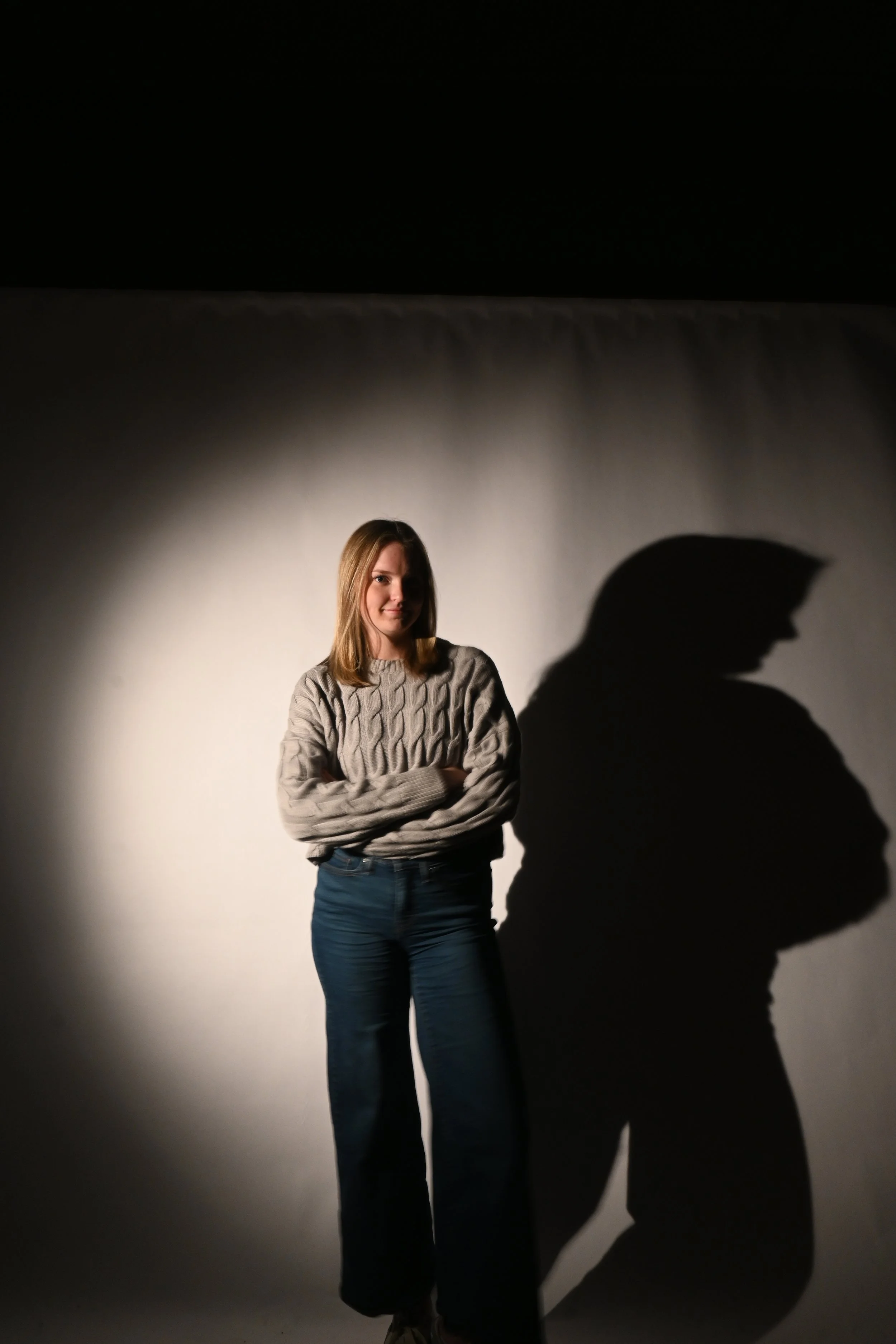 A young woman in a gray sweater and jeans standing with arms crossed, casting a large shadow on a plain wall.