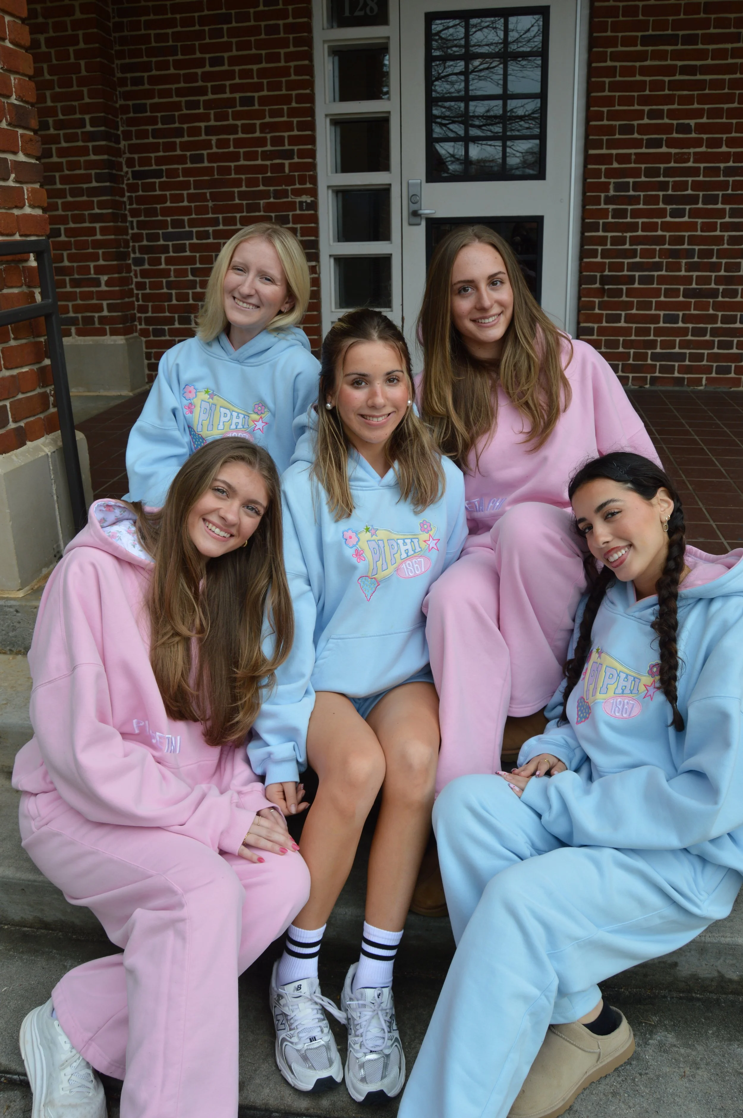 Group of five young women sitting on steps outside a brick building, wearing mostly pastel-colored hoodies and sweatpants, smiling at the camera.