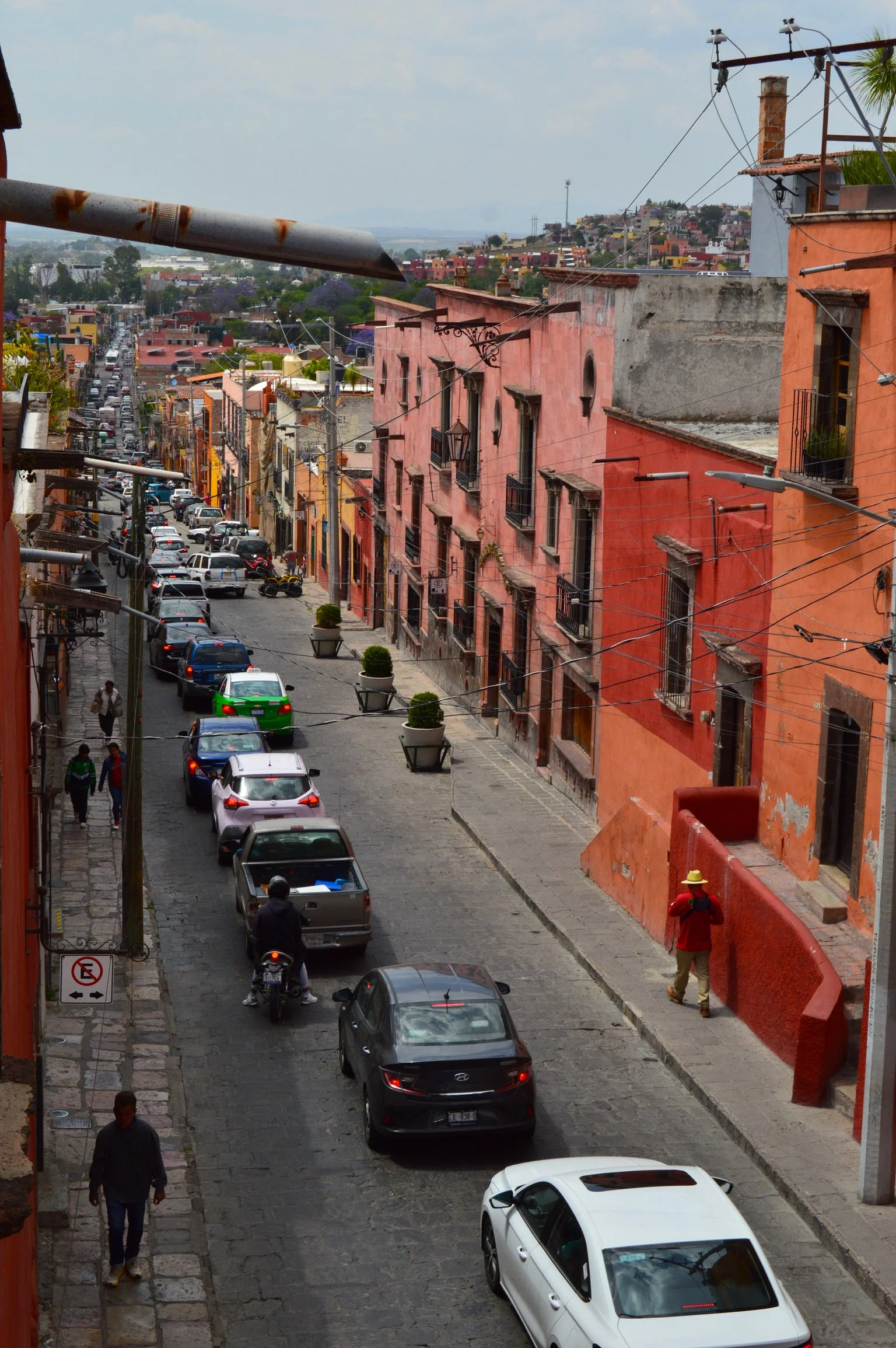 A narrow cobblestone street lined with colorful buildings, parked cars, pedestrians, and a person on a motorcycle, with utility wires overhead and a cityscape in the background.