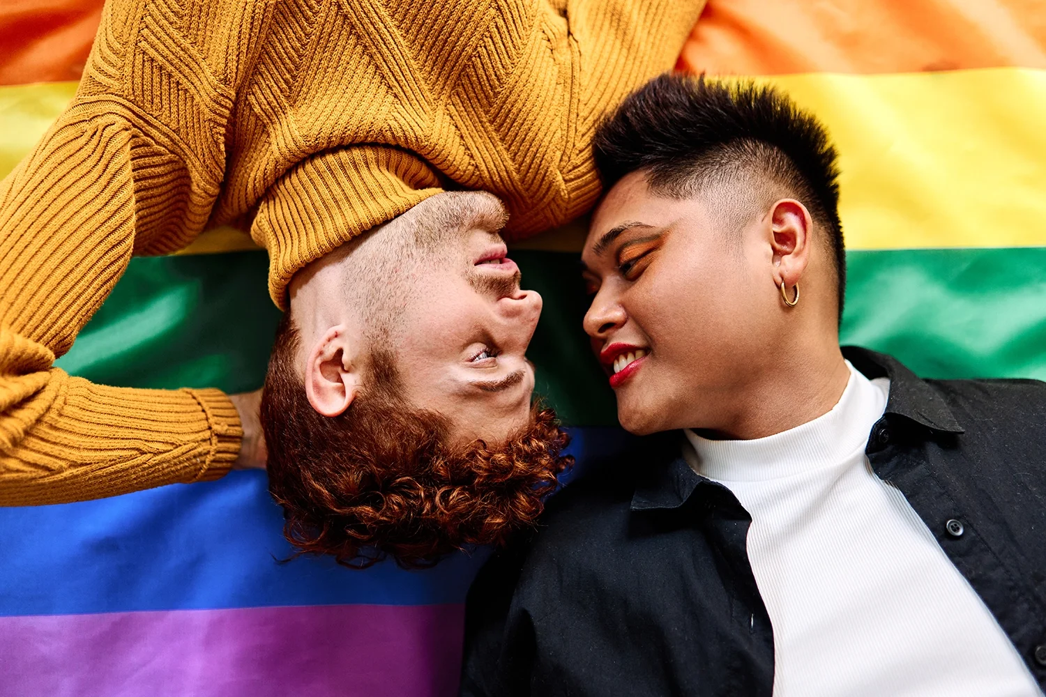 Two people lying on a rainbow pride flag, face to face, with eyes closed, smiling, in an intimate moment.