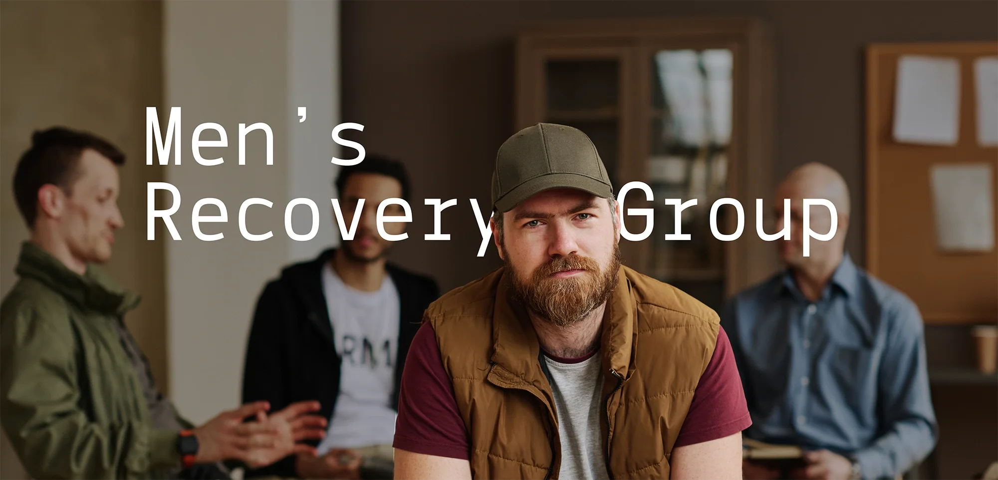 A man with a beard and wearing a baseball cap sitting in a support group meeting for men's recovery. Several other men are sitting behind him in a room with framed pictures on the wall.
