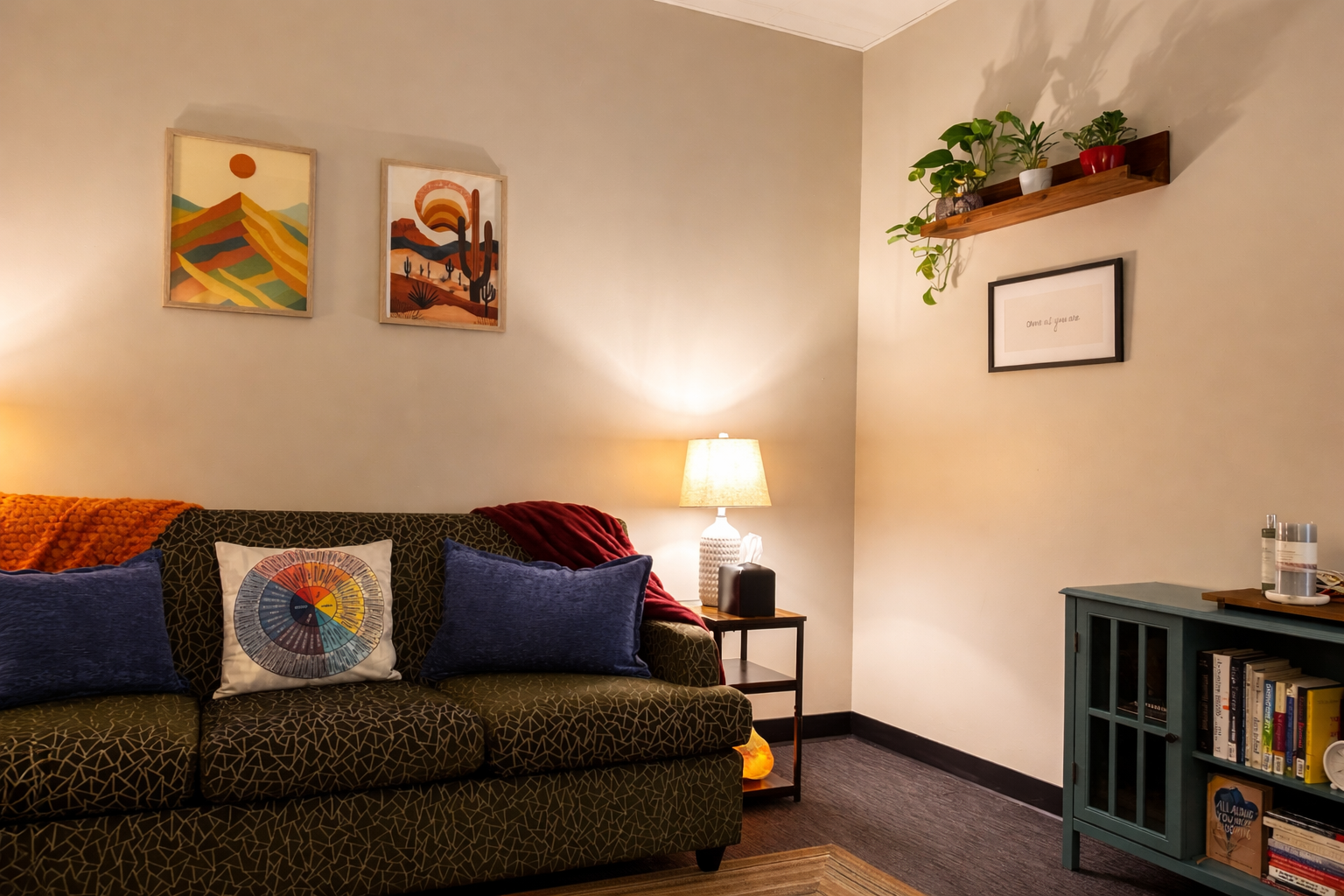 A cozy living room with a patterned dark green sofa adorned with colorful pillows, a side table with a lamp, artwork on the wall, potted plants on a wooden shelf, and a blue cabinet with books.