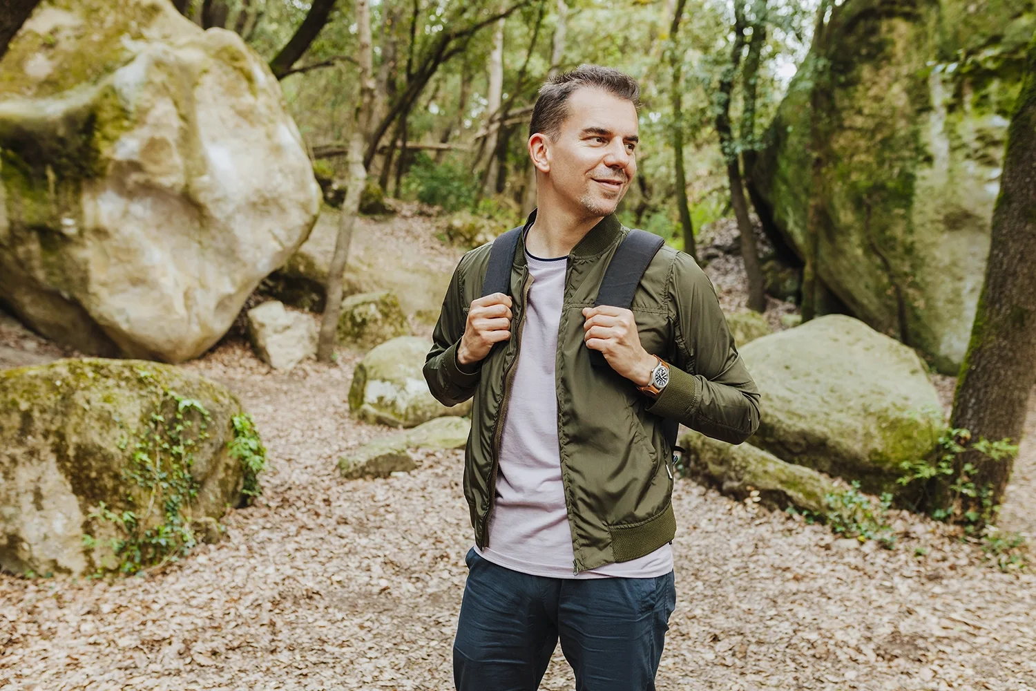 A man standing on a forest trail with foliage and large rocks around him, wearing a green jacket, a white t-shirt, dark pants, a backpack, and a watch, smiling and looking to the side.