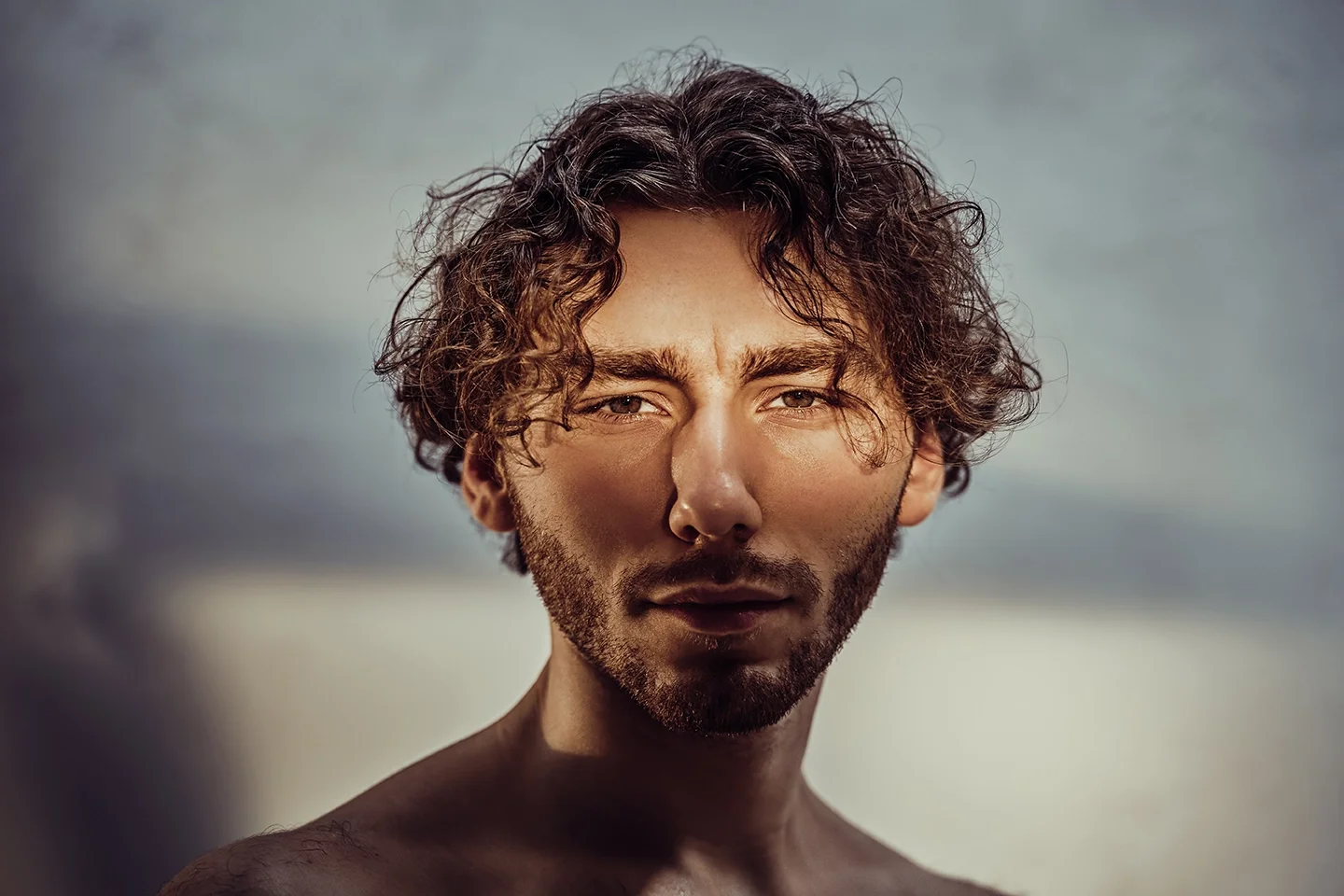 Close-up portrait of a young man with curly hair and facial hair, looking at the camera against a cloudy sky background.