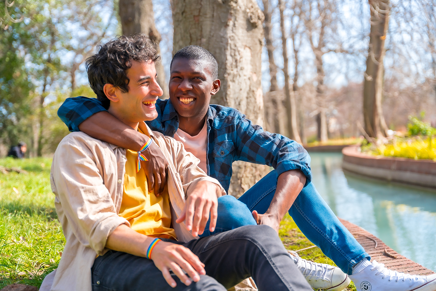 Two young men sitting by a canal, smiling and enjoying each other's company outdoors on a sunny day.