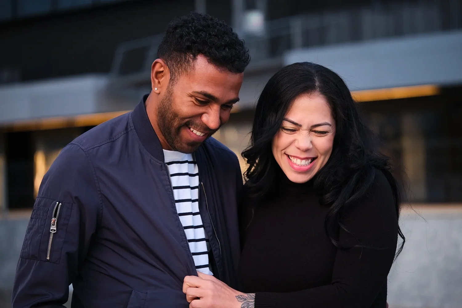 Two people smiling and laughing together outdoors, a man with short curly hair and a woman with long black hair, both dressed casually.