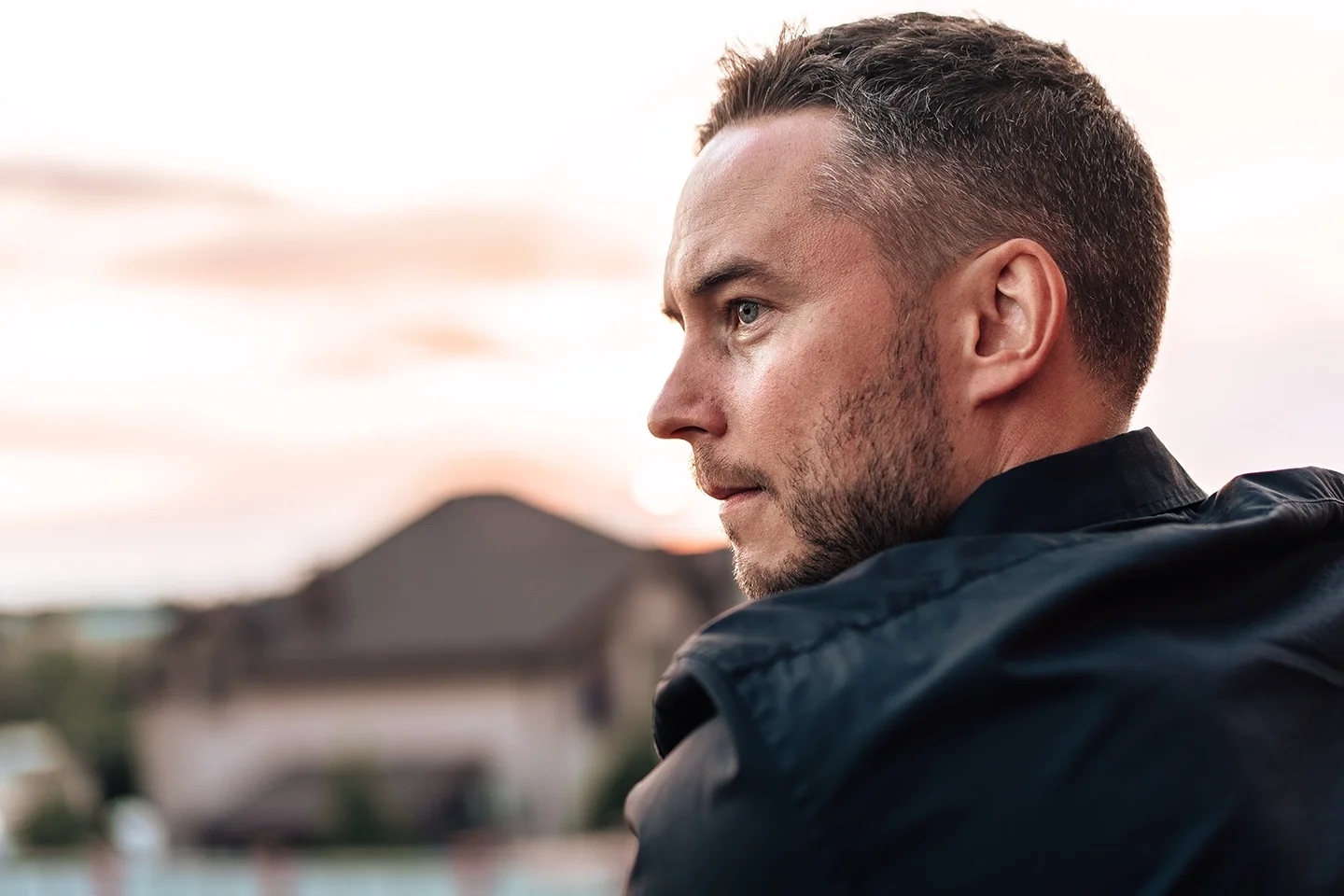 Profile of a man with short hair and a beard wearing a black jacket, gazing into the distance during sunset with houses blurred in the background.