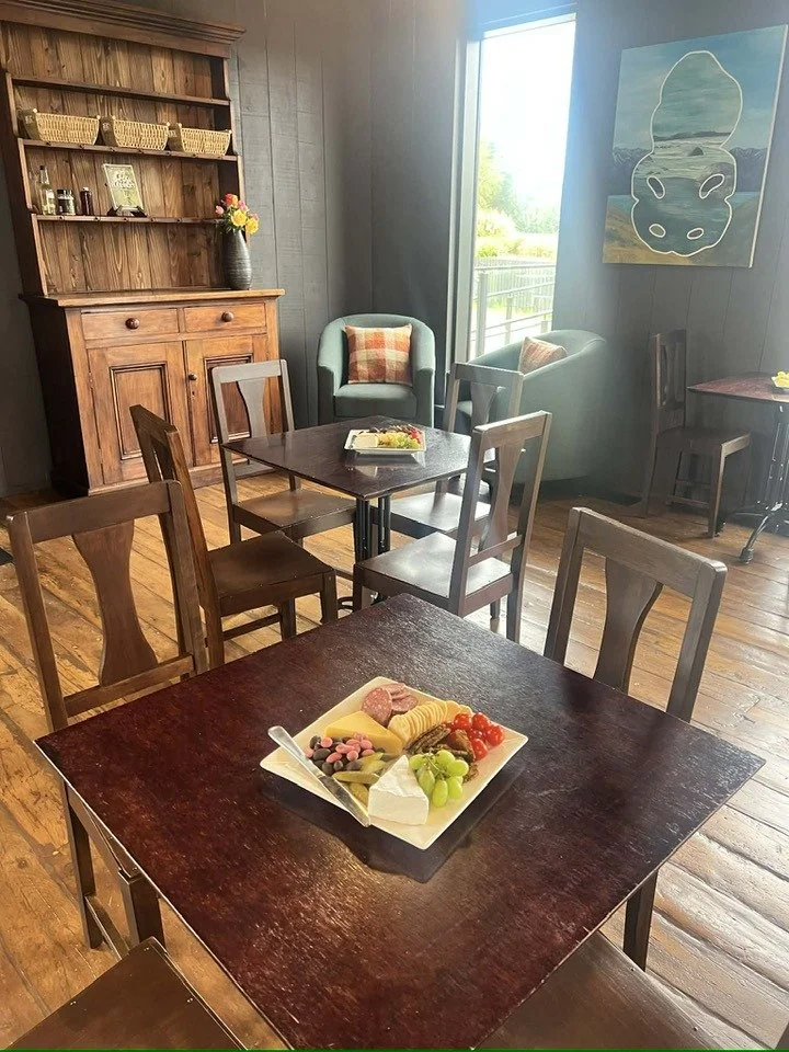 A cozy dining area with a wooden table set with a cheese and meat platter, surrounded by wooden chairs. In the background, there are two armchairs with pillows near a large window, a wooden hutch with baskets and decorative items, and a wall art piec