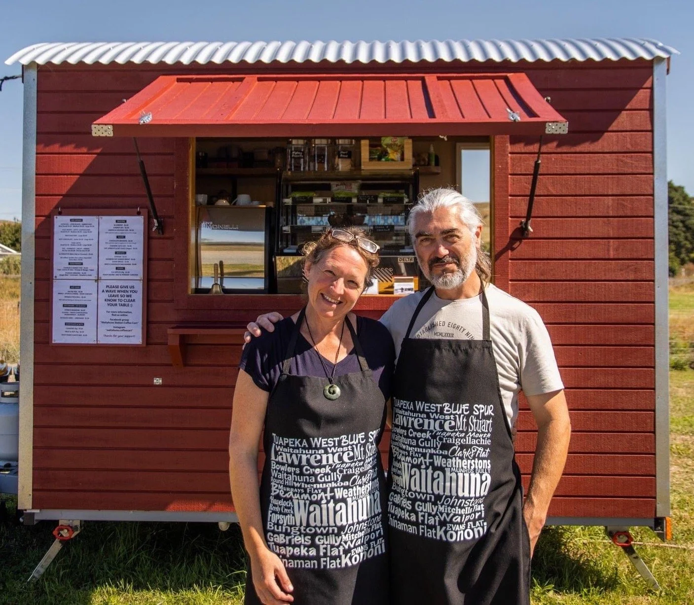 Two people standing in front of a small red food stand or kiosk, smiling and wearing black aprons with white text, outdoors on a sunny day.