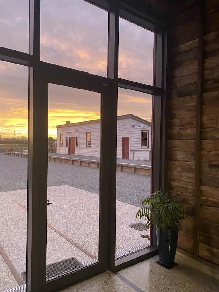 View through a glass door showing a small white building with wooden doors outside during sunset, with a gravel area and a wooden building with a sign reading 'Waitahiua'. Inside, there is a potted green plant near the right side of the door against 