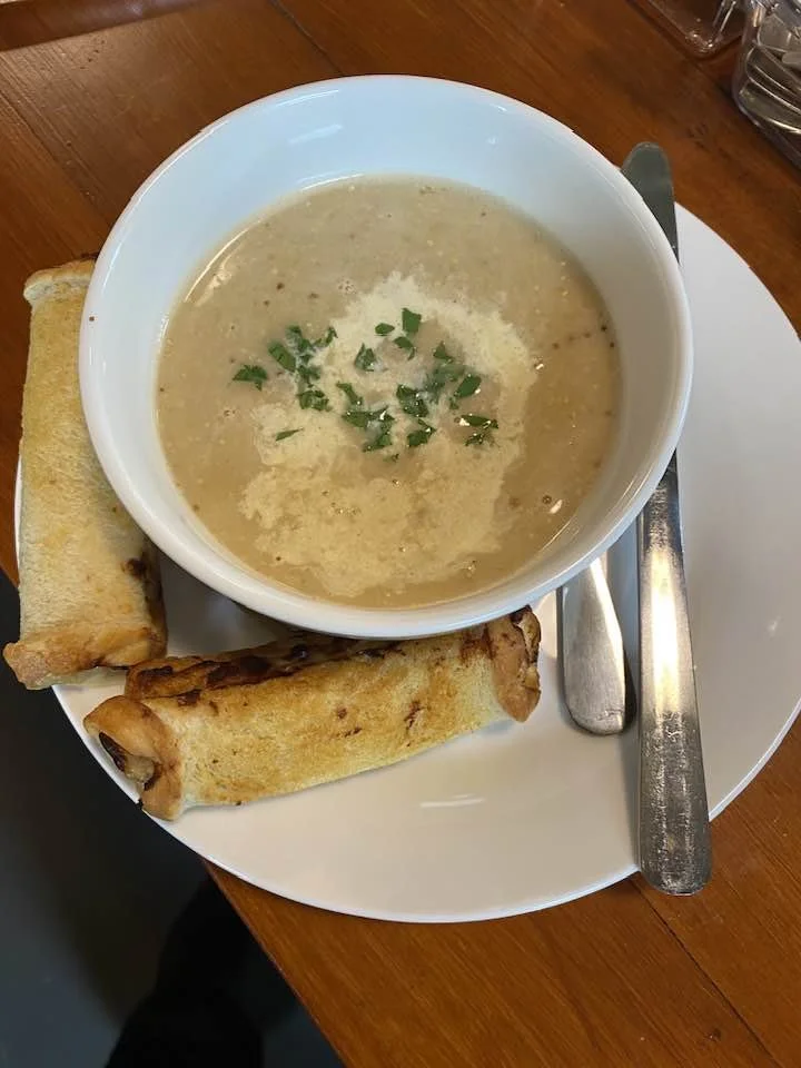 A bowl of creamy soup garnished with chopped herbs, served with two cheese rolls on a white plate, accompanied by a knife and fork on a wooden table.
