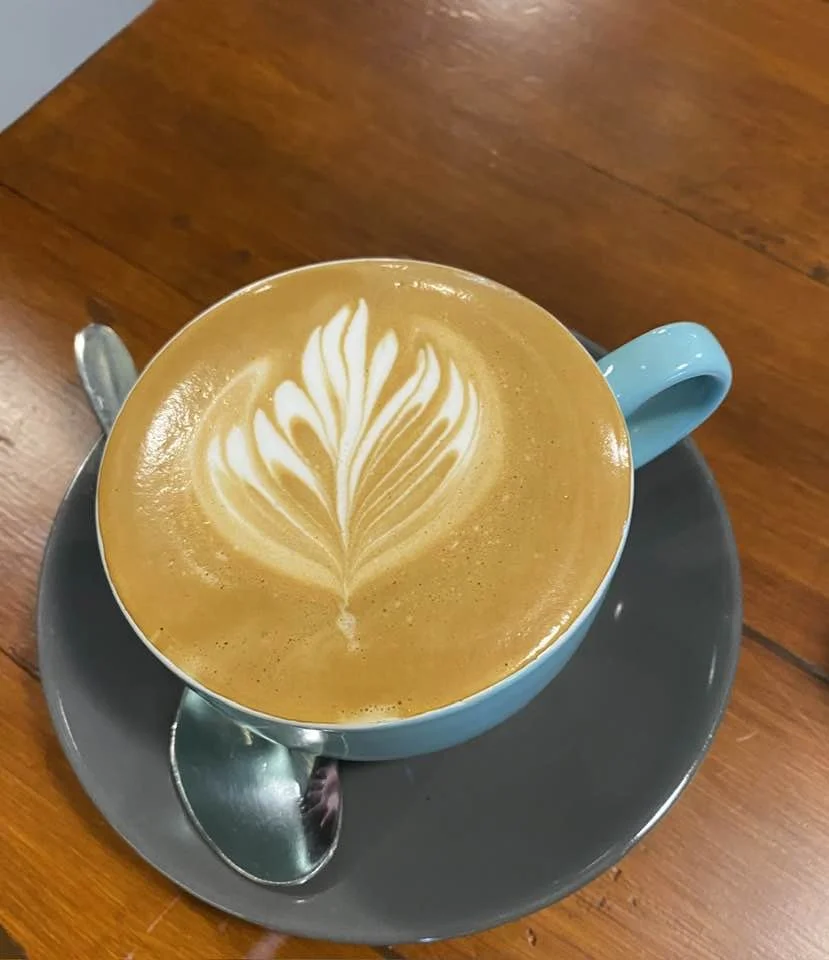 A blue coffee cup filled with a latte topped with latte art in the shape of a leafy feather design, sitting on a gray saucer with a silver spoon on a wooden table.