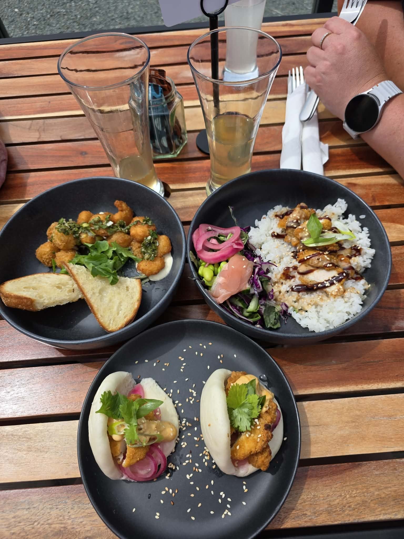 Two bowls of Asian cuisine on a wooden table with drinks and utensils. One bowl contains fried chicken pieces with green vegetables and toast, the other bowl has rice with vegetables, chicken, sauce, and pickled vegetables. There are three glasses of sparkling beverage.