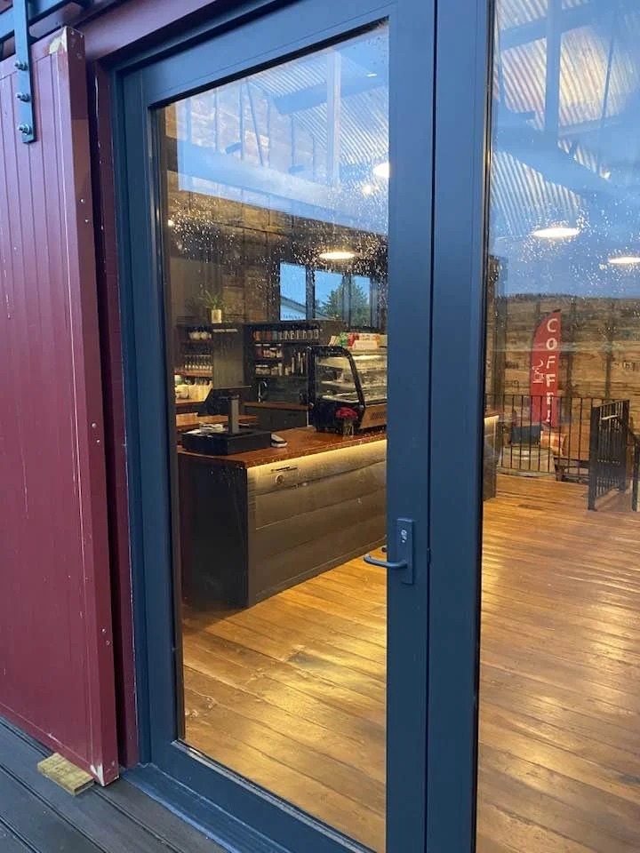 Interior of a Waitahuna Station Goods Shed Cafe viewed through a glass door, with a counter, coffee machine, and seating area faintly visible.