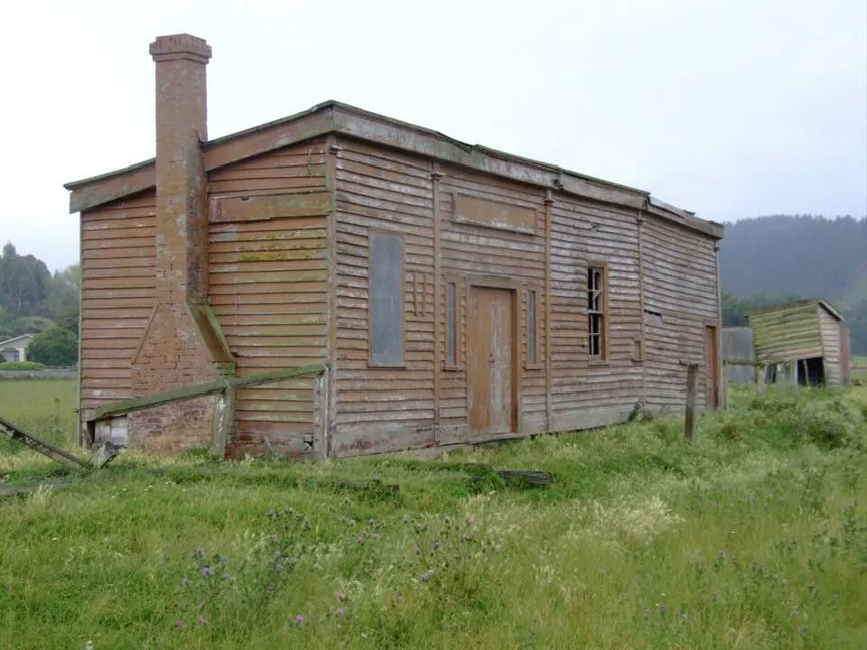 An old, weathered wooden house on a grassy field with a hill in the background, showing signs of disrepair.