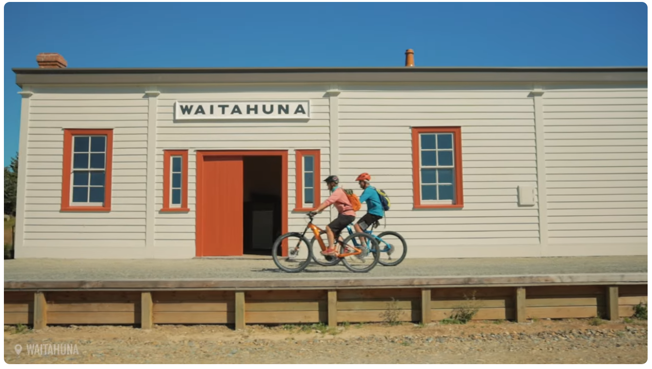 Two people riding bicycles in front of a building with a sign that reads 'WAITAHUNA'. The building has white siding with orange trim around the windows and door, and a blue sky overhead.