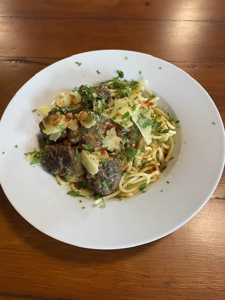 Plate of spaghetti topped with meatballs, grated cheese, chopped herbs, and a sprinkle of red spice, on a wooden table.