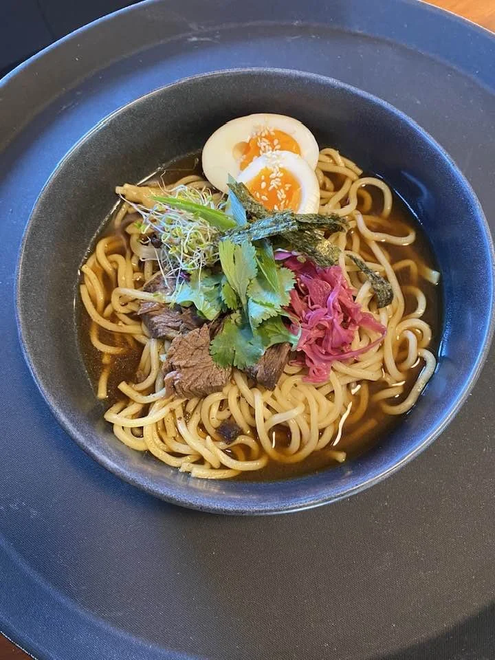 Bowl of ramen noodles with sliced beef, a soft-boiled egg, seaweed, green herbs, microgreens, and pickled ginger.