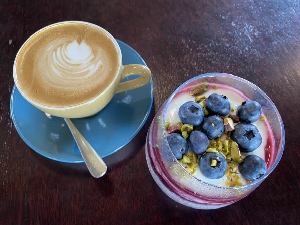 A cup of coffee with latte art next to a chai bowl of yogurt topped with blueberries and chopped pistachios on a dark wooden table.
