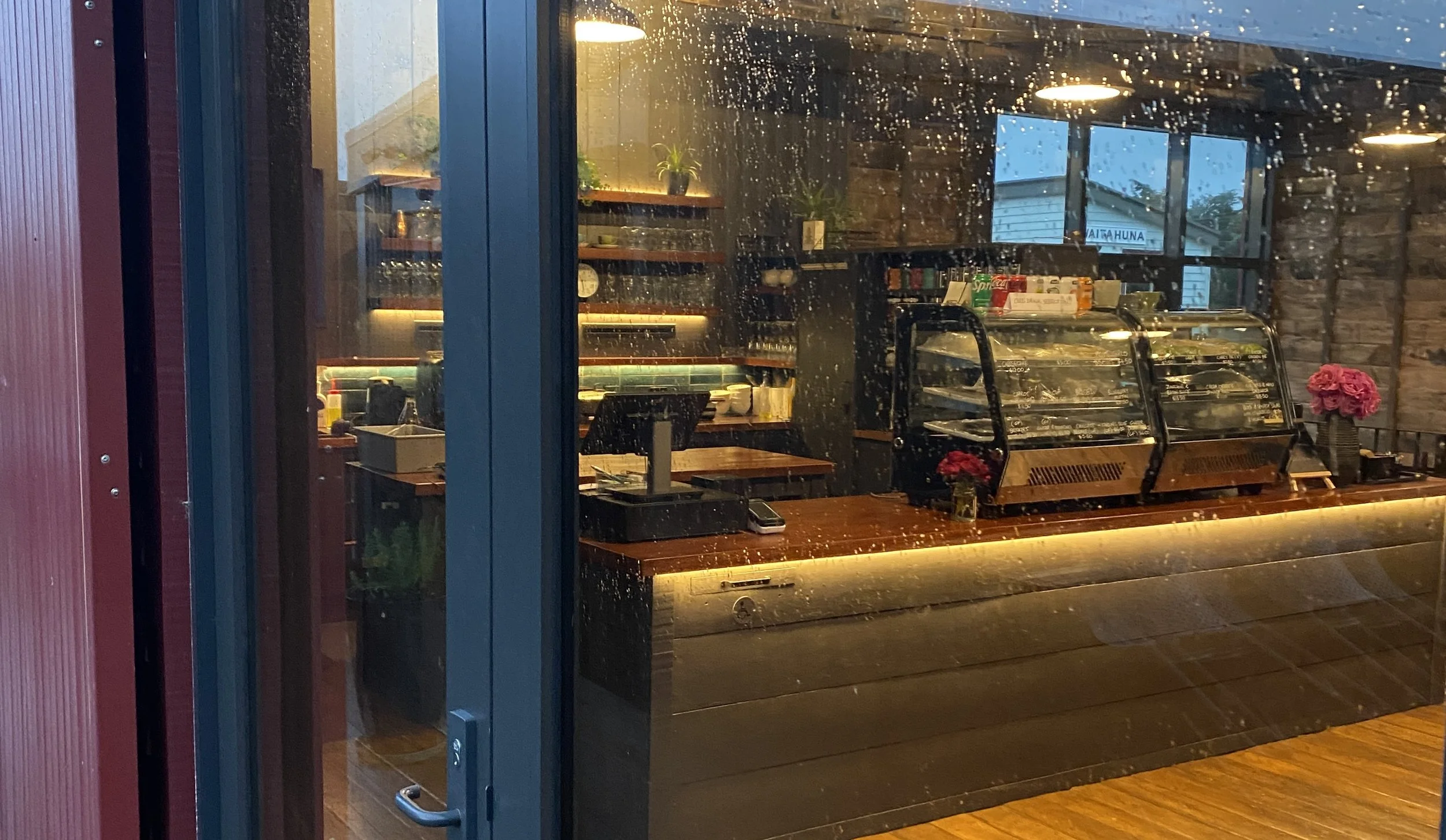 View of a restaurant or cafe interior from outside through a rain-streaked glass window, showing a counter with a glass display case, pink flowers, and shelves with dishes and cups, illuminated with warm lighting.