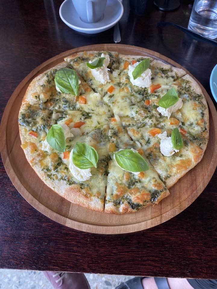 A pizza topped with cheese, fresh basil leaves, dollops of ricotta cheese, and small pieces of orange, green, and yellow vegetables on a wooden serving tray.