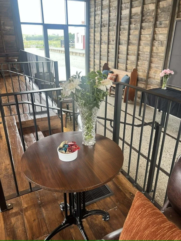 Waitahuna Station Goods Shed Cafe. A small round wooden table with a vase of white daisies and a bowl of wrapped candies. In the background, there is a cushioned bench with pillows, a high glass window letting in natural light.