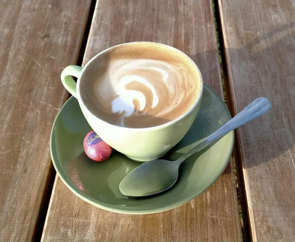 A cup of coffee with foam art, placed on a green saucer with a spoon and a small easter egg, on a wooden table.