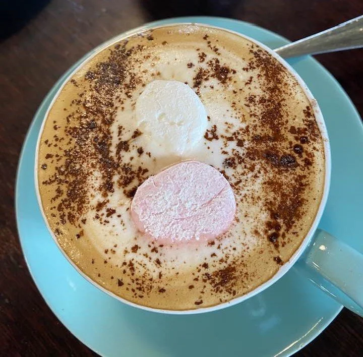 A cup of cappuccino topped with whipped cream, chocolate sprinkles, and marshmallows on a blue saucer with a spoon.