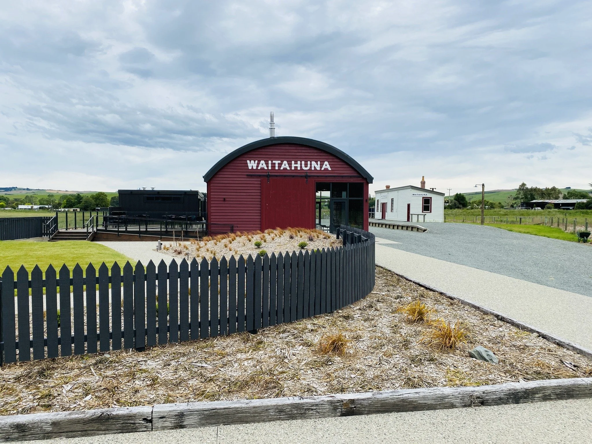 Waitahuna Station Goods Shed Cafe, maroon building situated on a gravel area with a black fence and a small garden. There is a white building with the same name in the background, and green hills with a cloudy sky overhead.
