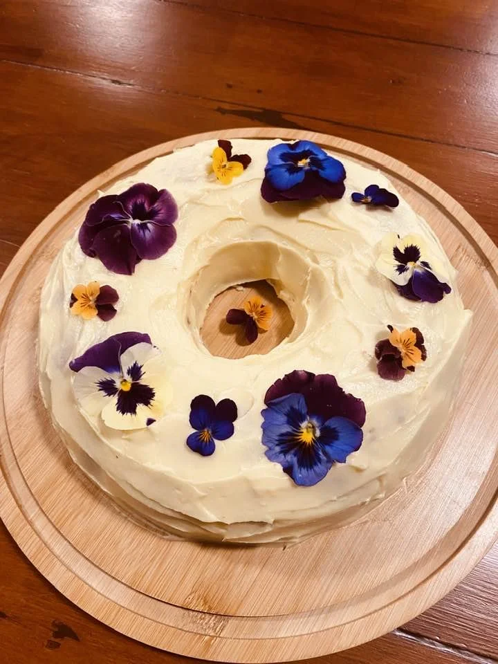 A round cake with cream frosting decorated with colorful edible pansy flowers on top. The cake is on a wooden serving board placed on a wooden table.