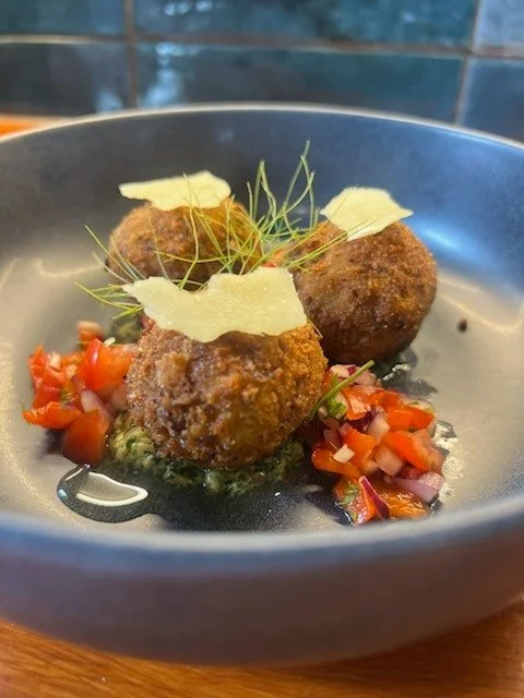 Three fried arancini garnished with shaved Parmesan cheese and microgreens, served with chopped tomatoes on a black plate.