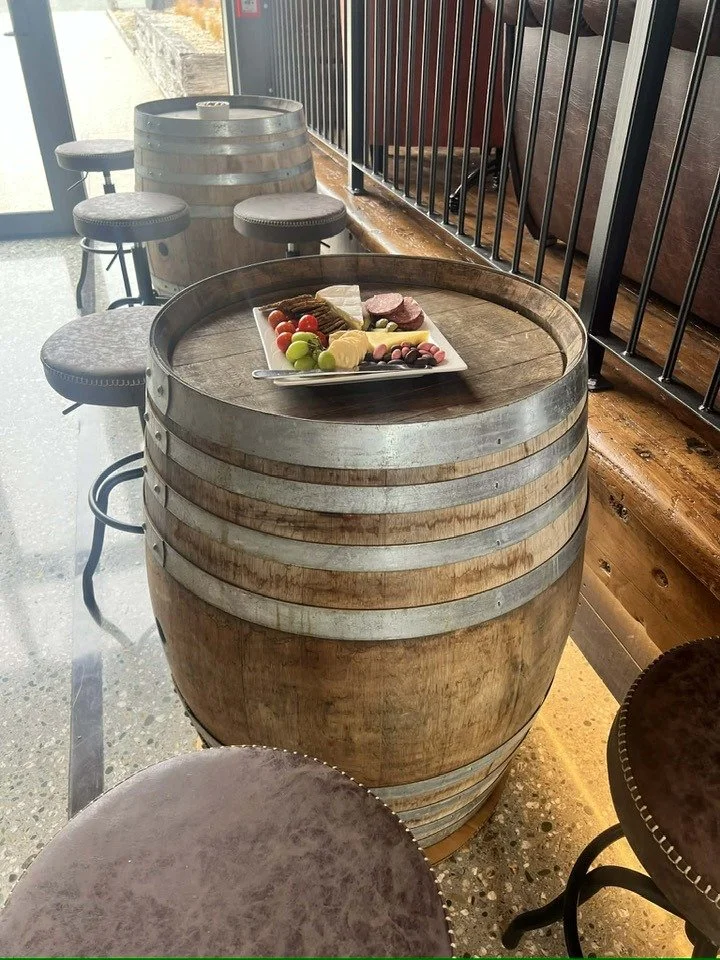 Wine barrel table with a platter of cheese, grapes, crackers, and meats, surrounded by bar stools inside a restaurant.