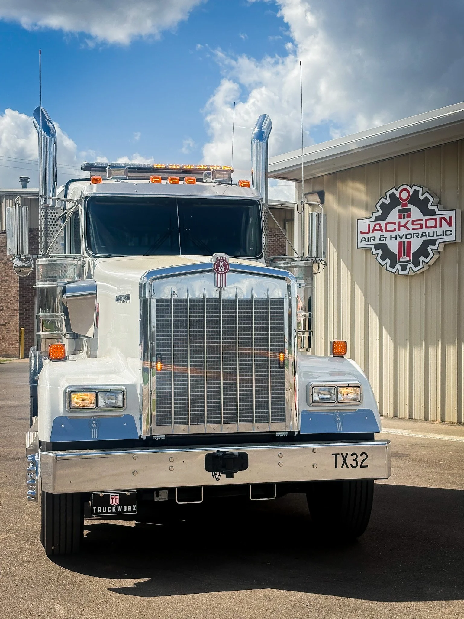 Front view of a large white semi-truck parked outdoors in front of a building with a sign for Jackson Air & Hydraulic. The truck has chrome accents, tall exhaust pipes, and a Texas license plate TX32.