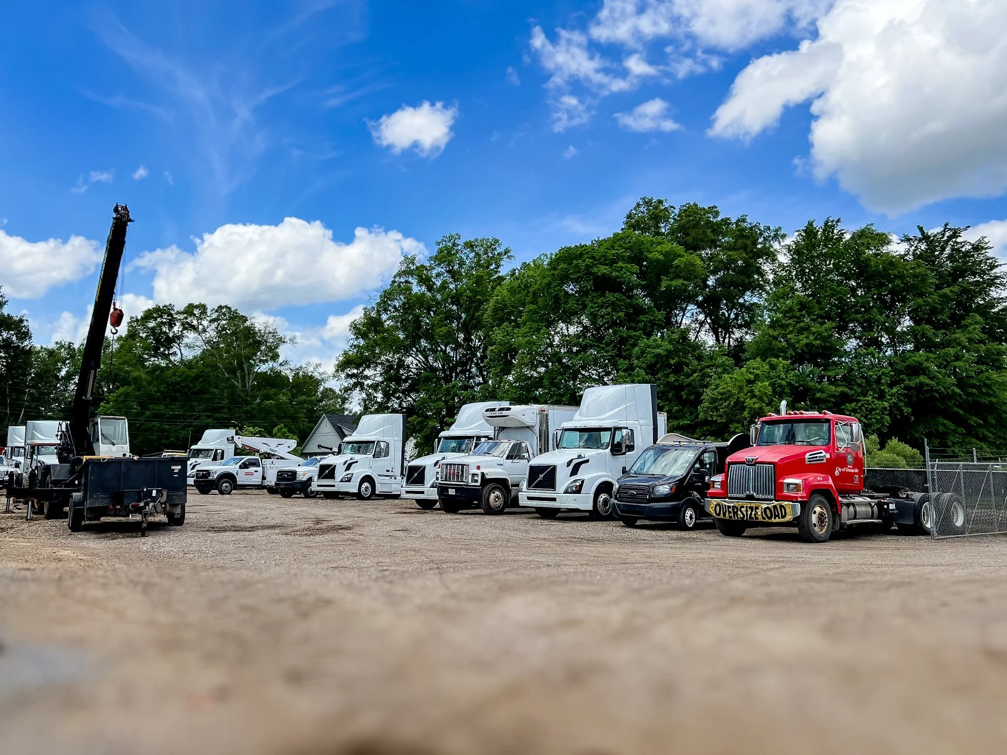 A parking lot filled with various trucks, including semi-trucks and utility vehicles, some marked with company logos, with a backdrop of green trees and a partly cloudy blue sky.