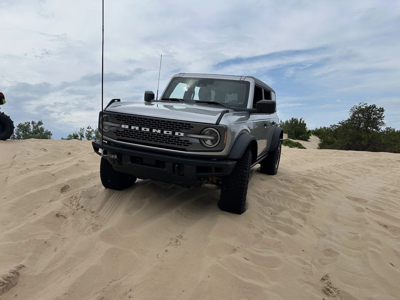 A gray Ford Bronco off-road vehicle parked on sandy terrain with a partly cloudy sky and some trees in the background.