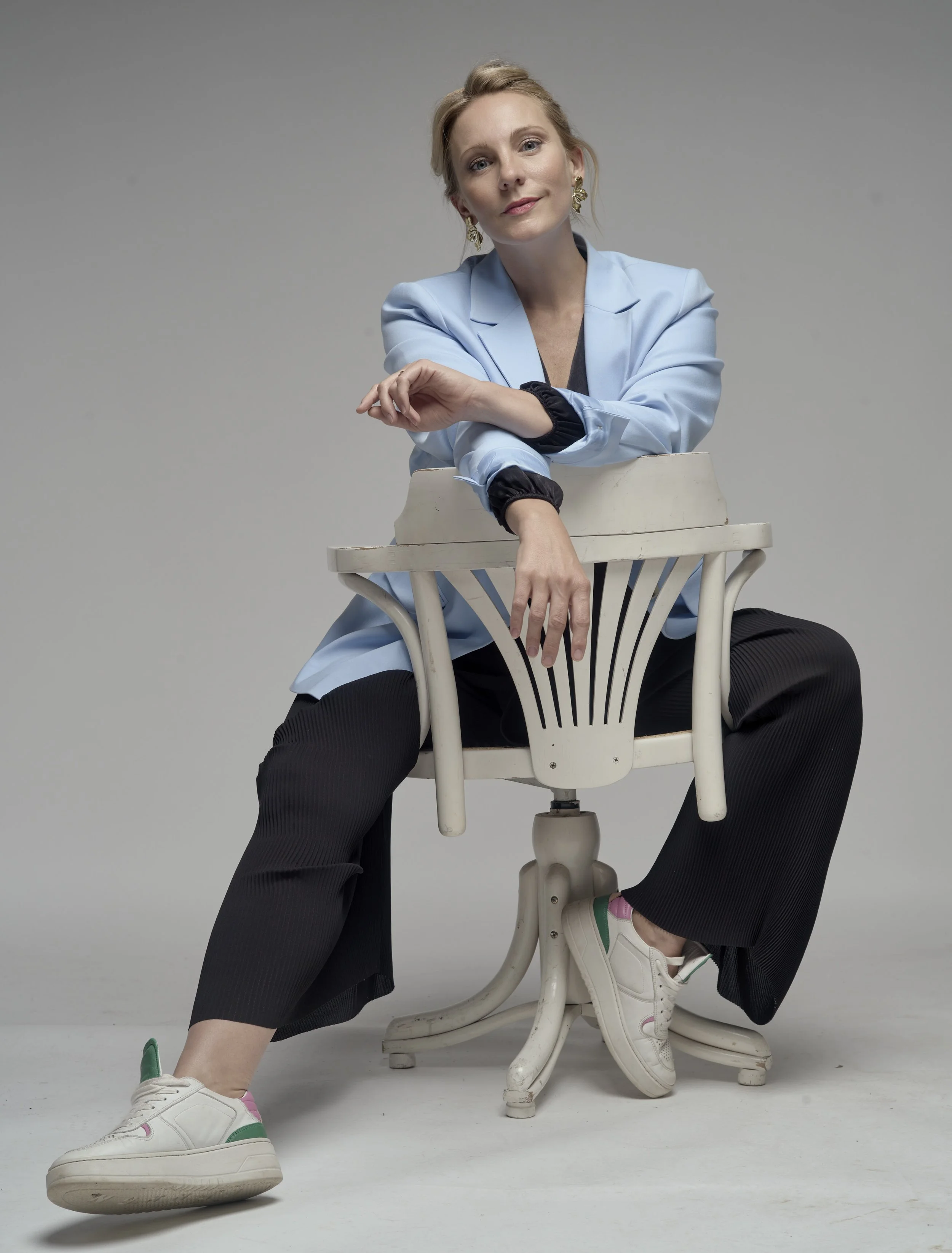Eine Frau sitzt auf einem weißen Stuhl, trägt einen hellblauen Blazer und schwarze Hosen, trägt weiße Sneakers mit grünen und pinken Akzenten, und hat blonde Haare. Sie sitzt entspannt, bewegt ein Bein, und blickt freundlich in die Kamera.