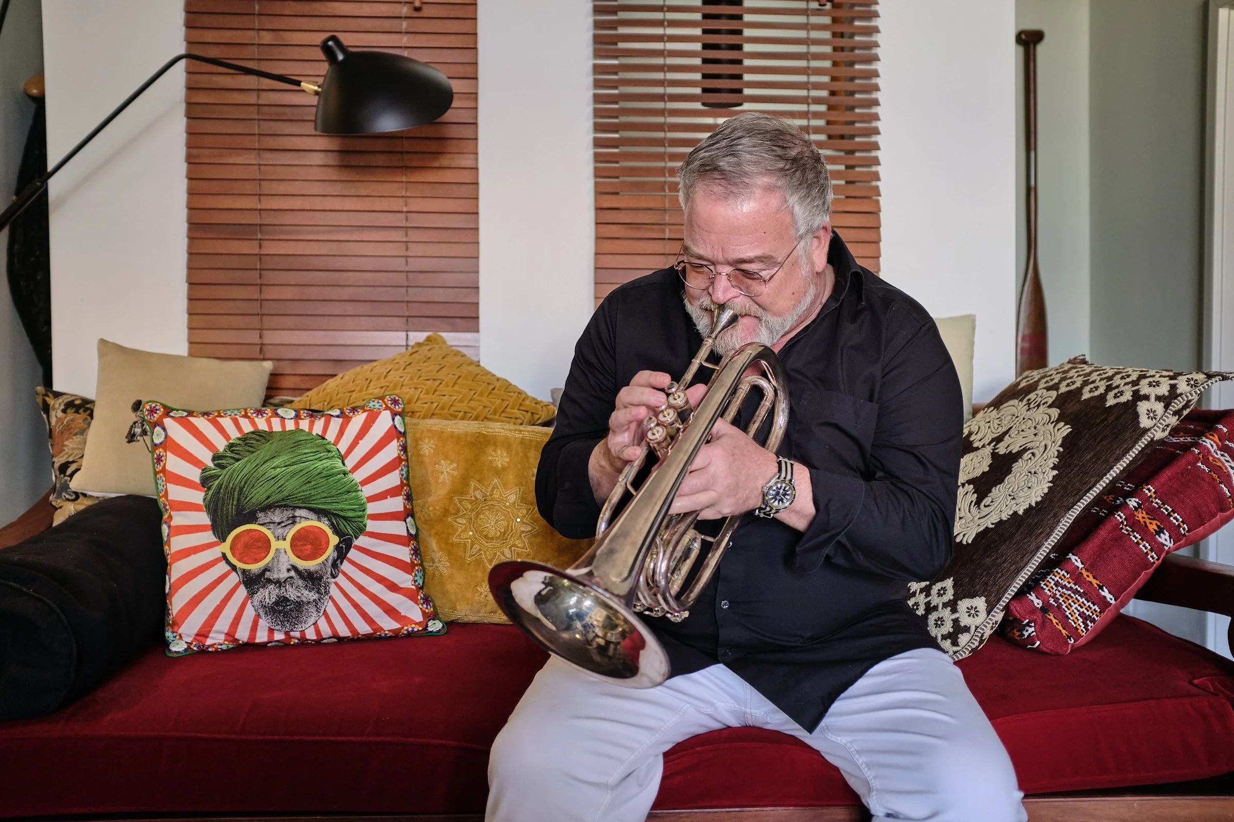 Ein älterer Mann mit grauem Haar und Brille spielt saxophon in einem Wohnzimmer mit bunten Kissen und Holzhintergrund.