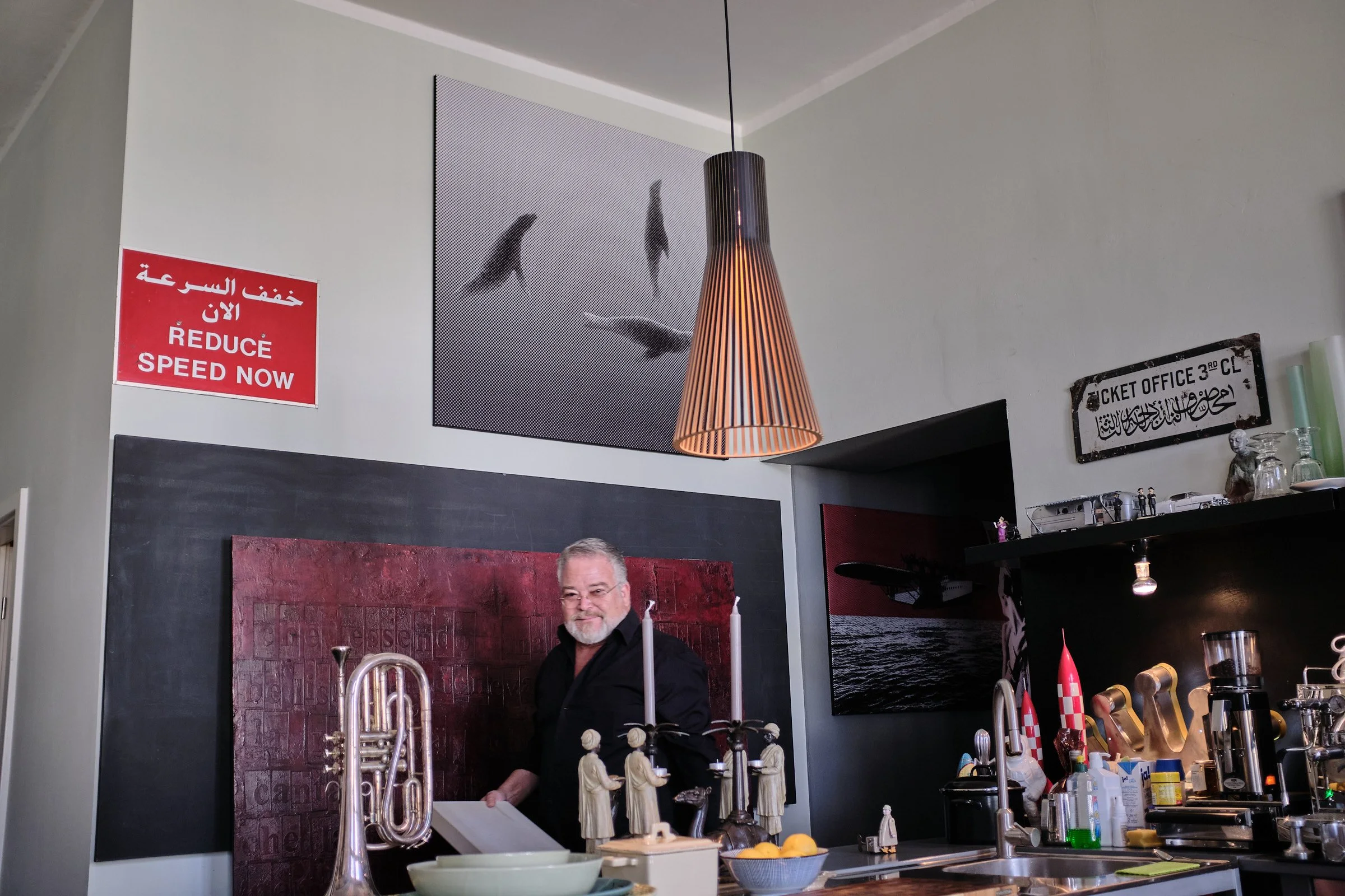 Ein Mann mit Brille und grauem Bart steht in einer Bar oder einem Restaurant, im Vordergrund sind Geschirr und Dekorationen sichtbar, im Hintergrund an der Wand sind Schilder mit Texten in verschiedenen Sprachen und an der Decke eine hängende Lampe.