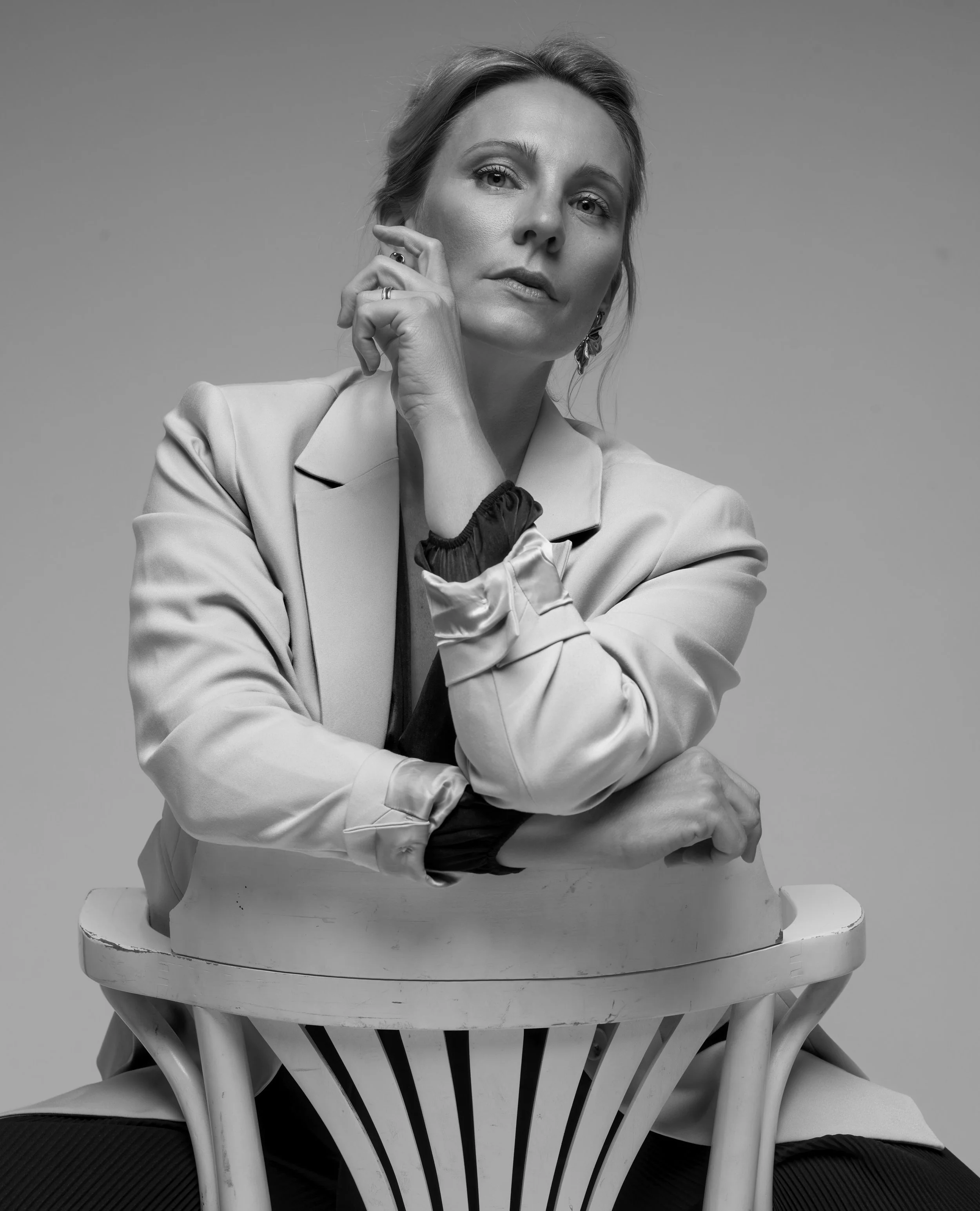Schwarz-weiß-Fotografie eines Model Femmes, sitzend an einem runden Stuhl, die Hand an ihrem Gesicht, mit einer nachdenklichen Mimik, elegante Kleidung und Schmuck, monochrome Ästhetik
