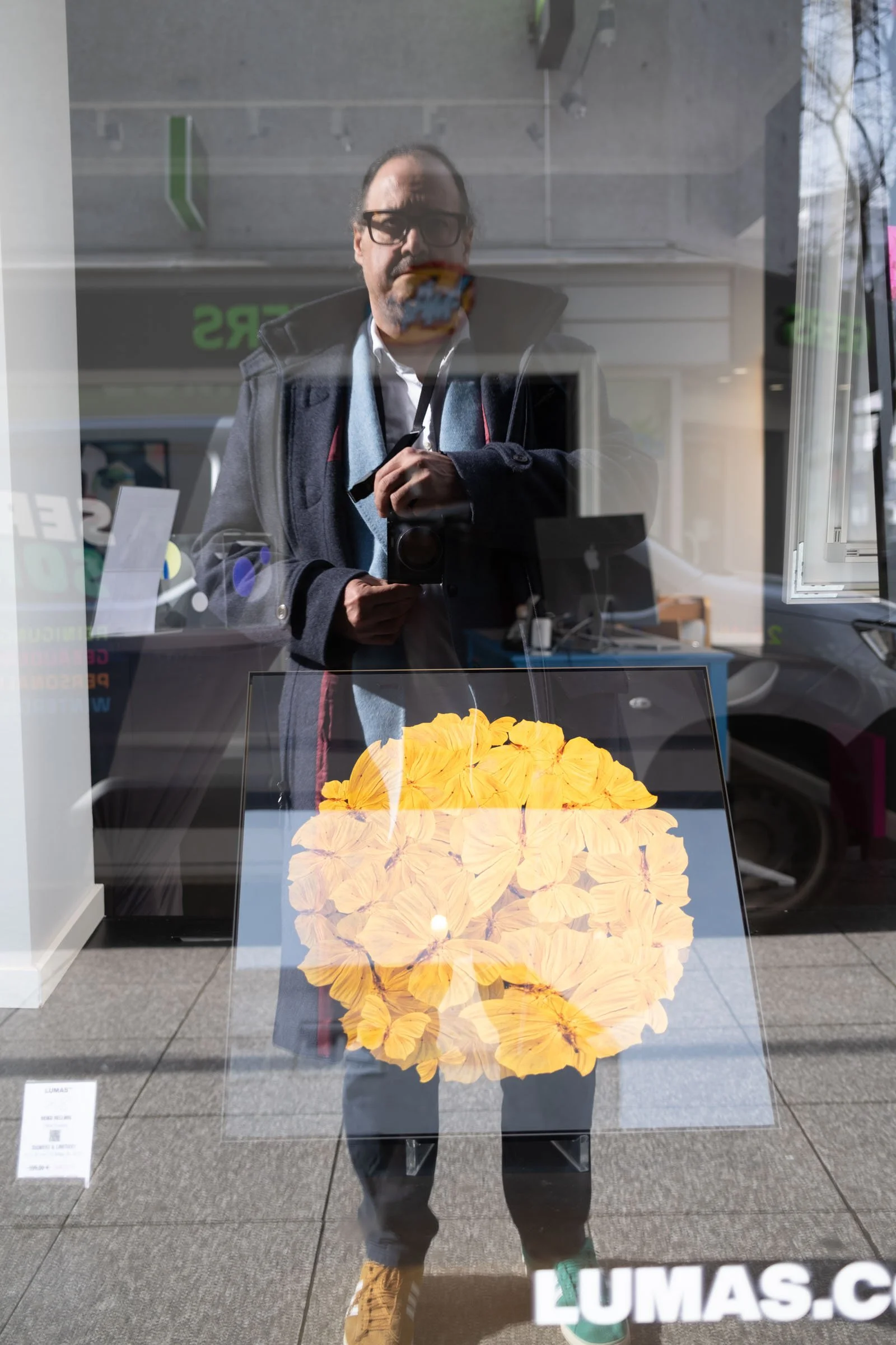 Reflexion eines Mannes mit Brille, dunklem Mantel und grauem Schal, der mit einer Kamera vor einem Schaufenster steht. Im Schaufenster ist ein gelbliches, blumenähnliches Dekor zu sehen, und die Umgebung spiegelt sich in der Scheibe.