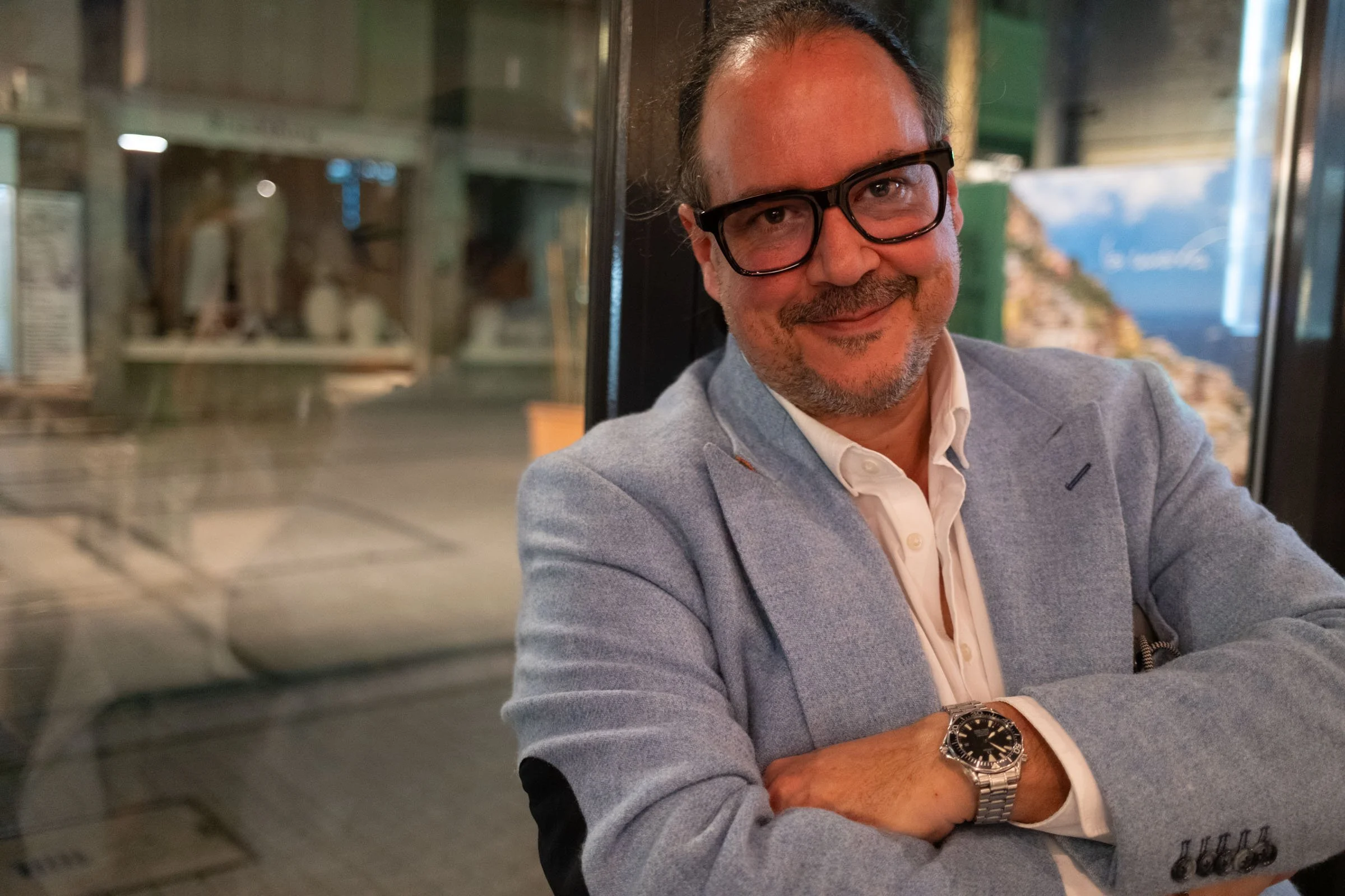 Ein lächelnder Mann mit Brille, grauem Blazer und weißem Hemd sitzt in einem Raum mit Glasfenstern und einem Wasserfall im Hintergrund.