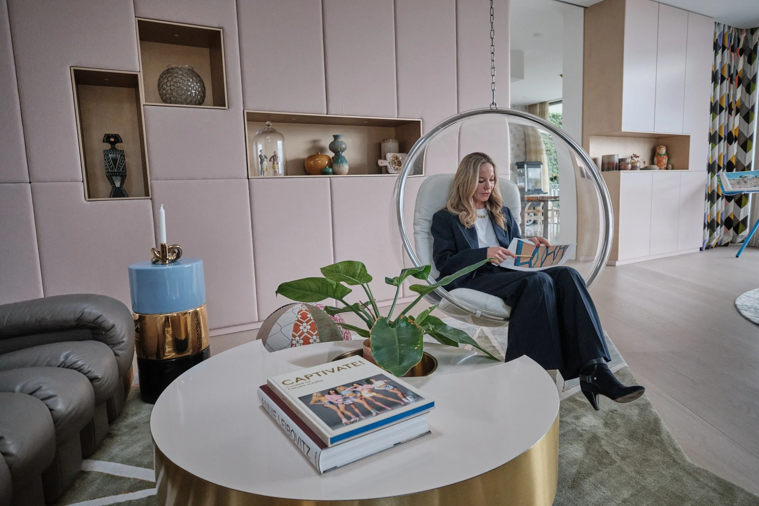 Frau sitzt in einem modernen Wohnzimmer in einem transparenten Sitzsessel, liest ein Buch. Im Vordergrund ein runder Couchtisch mit einem großen Buch namens "CAPTIVATE!", einer Pflanze und anderen Dekorationsgegenständen. Im Hintergrund eine Wand mit