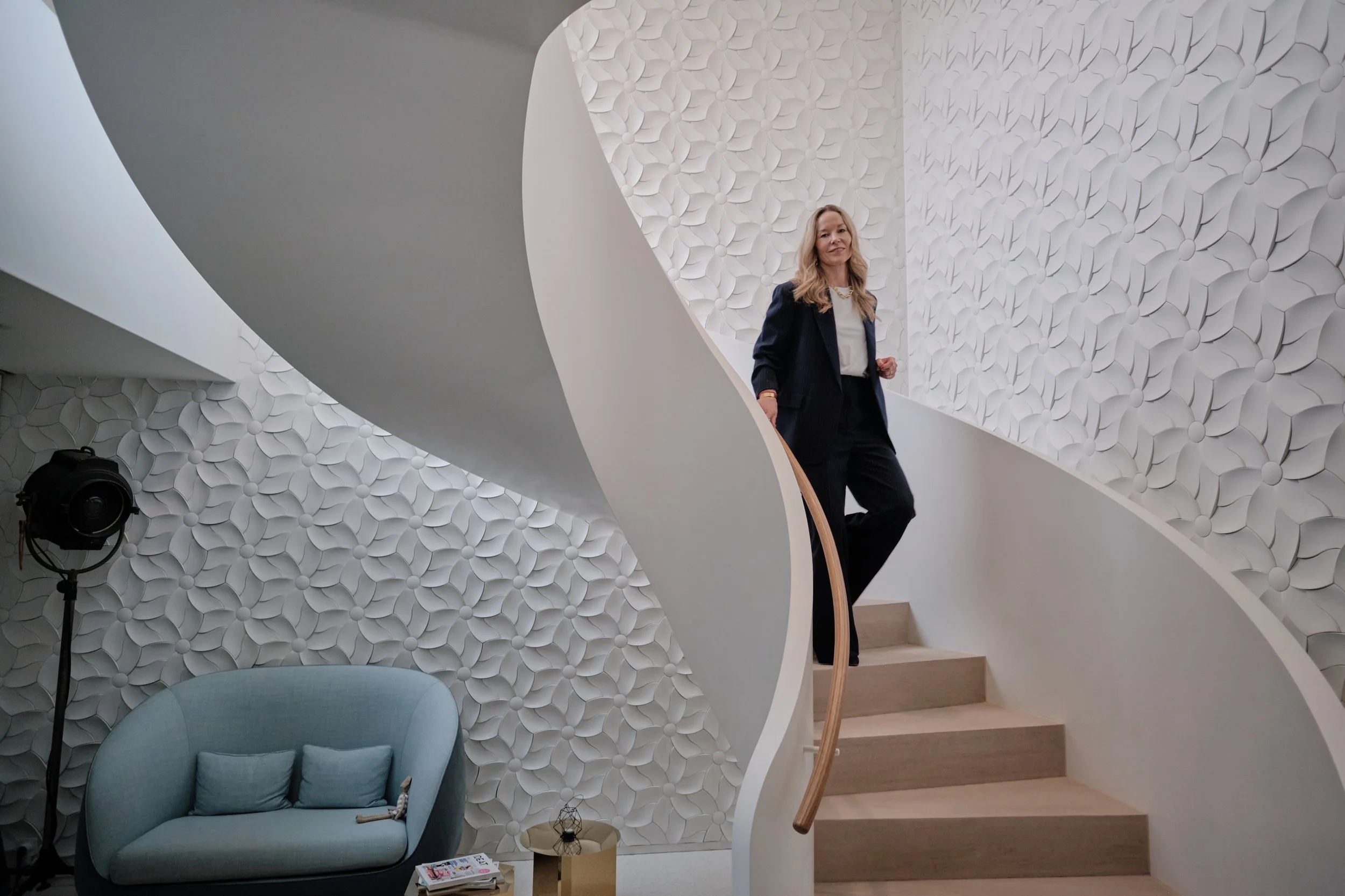 Frau in dunkler Kleidung steht an einer Treppe in einem stilvollen Raum mit weißen, blumenartig gestalteten Wandpaneelen.