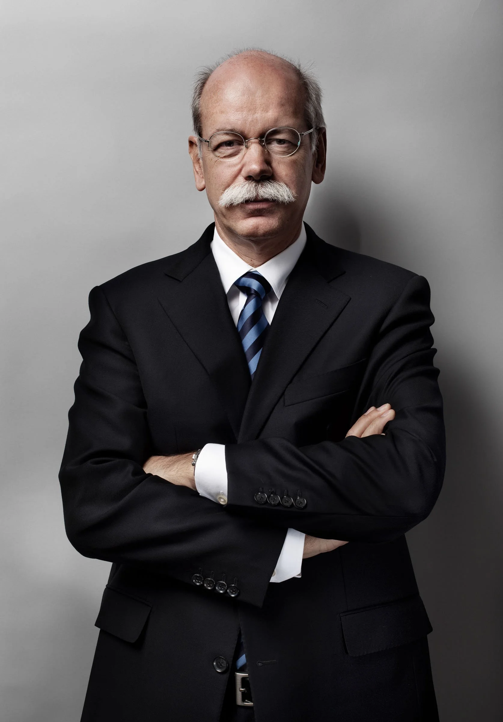 Ein älter Mann mit Brille, weißem Schnurrbart und Anzug steht vor einem grauen Hintergrund. Er hat die Arme verschränkt.