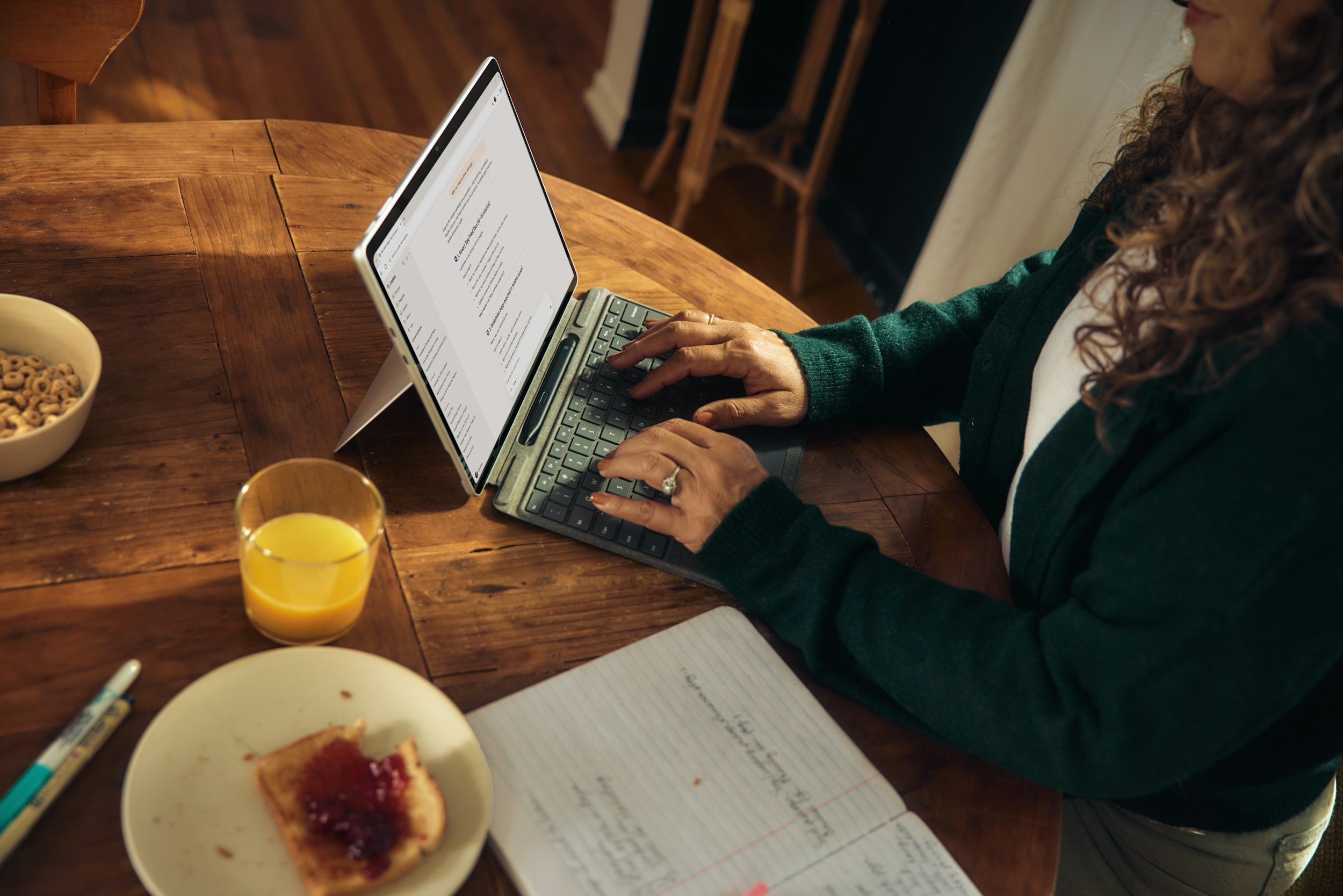 A person working on a tablet at a wooden table, with a notebook, a glass of orange juice, a bowl of cereal, a plate with a partially eaten piece of toast with jam, and a pen nearby.